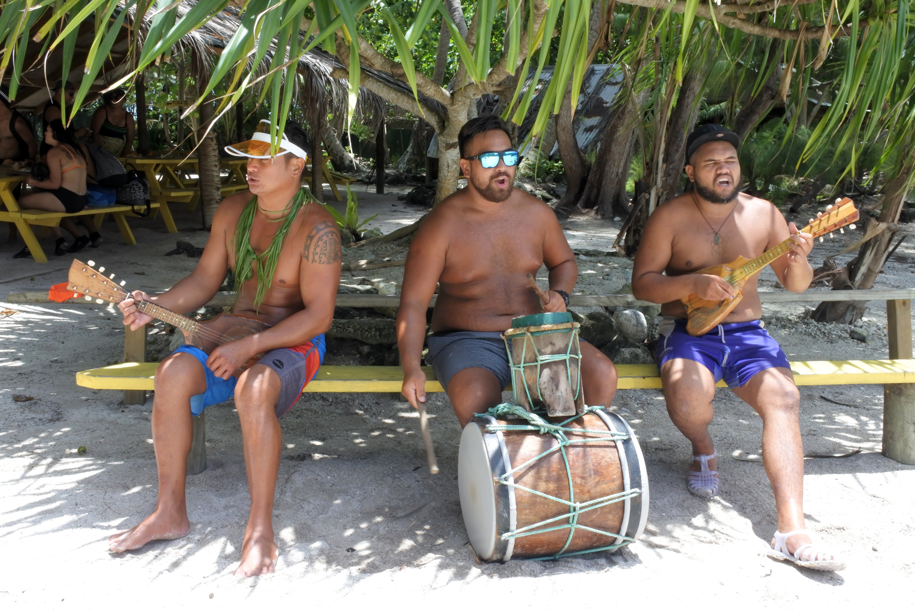 Three shirtless men play traditional instruments, with palm trees and a sandy beach in the background.