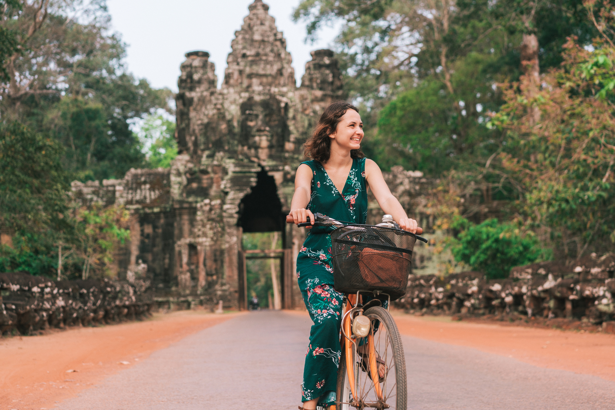 A woman rides a bicycle on a path near a historic stone temple surrounded by lush greenery.