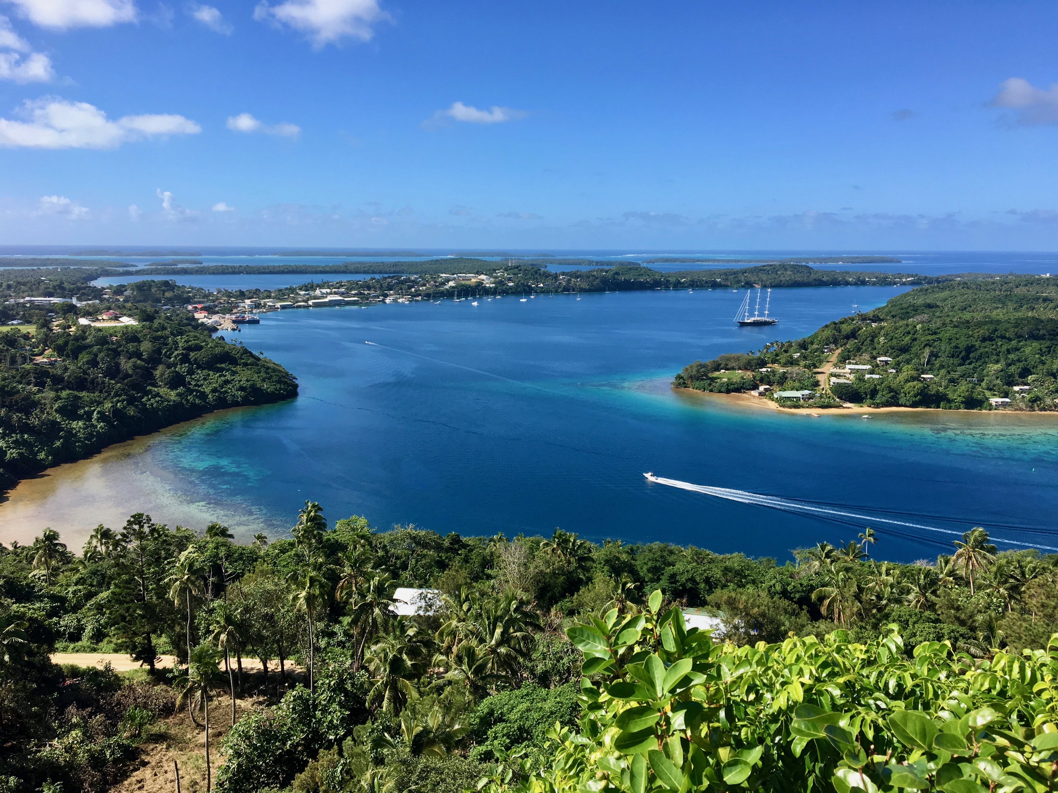 A scenic view of a bay with clear blue water, surrounded by greenery and small boats, under a bright blue sky.