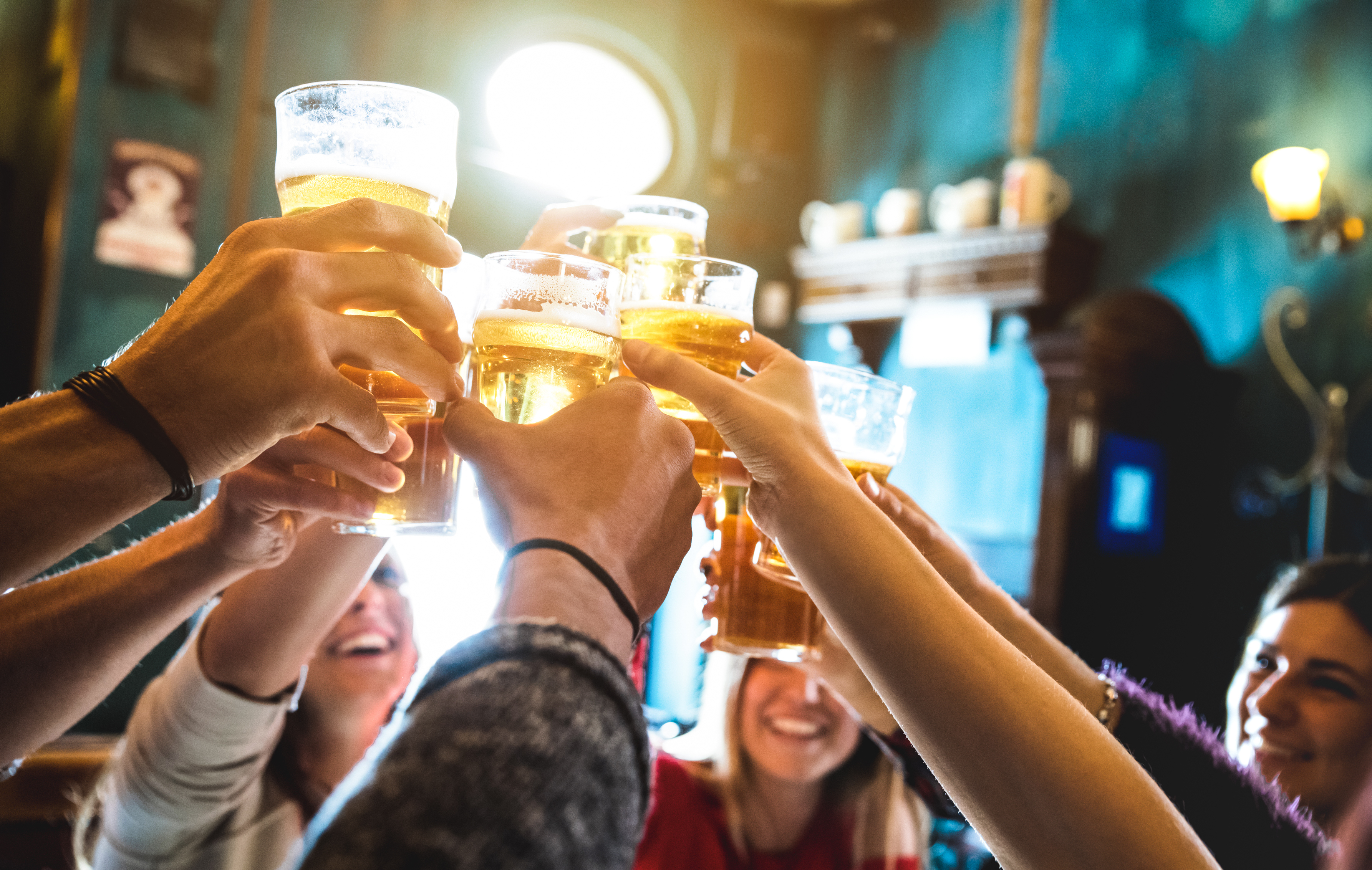 Group of people raising glasses of beer in a toast, smiling, inside a lively pub with colorful decor.