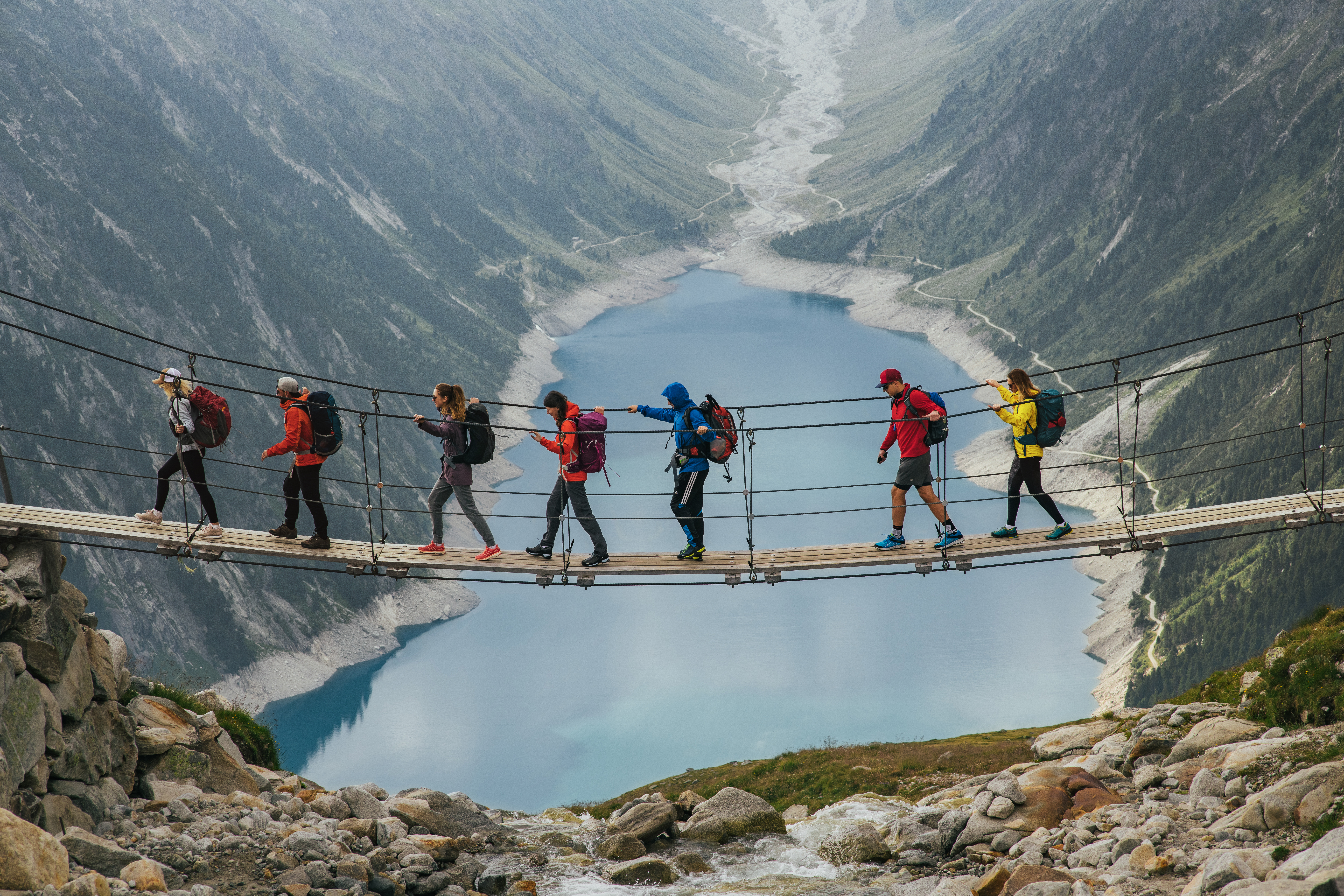  A group of travelers crosses a suspension bridge against the backdrop of a mountain and a glacier. 