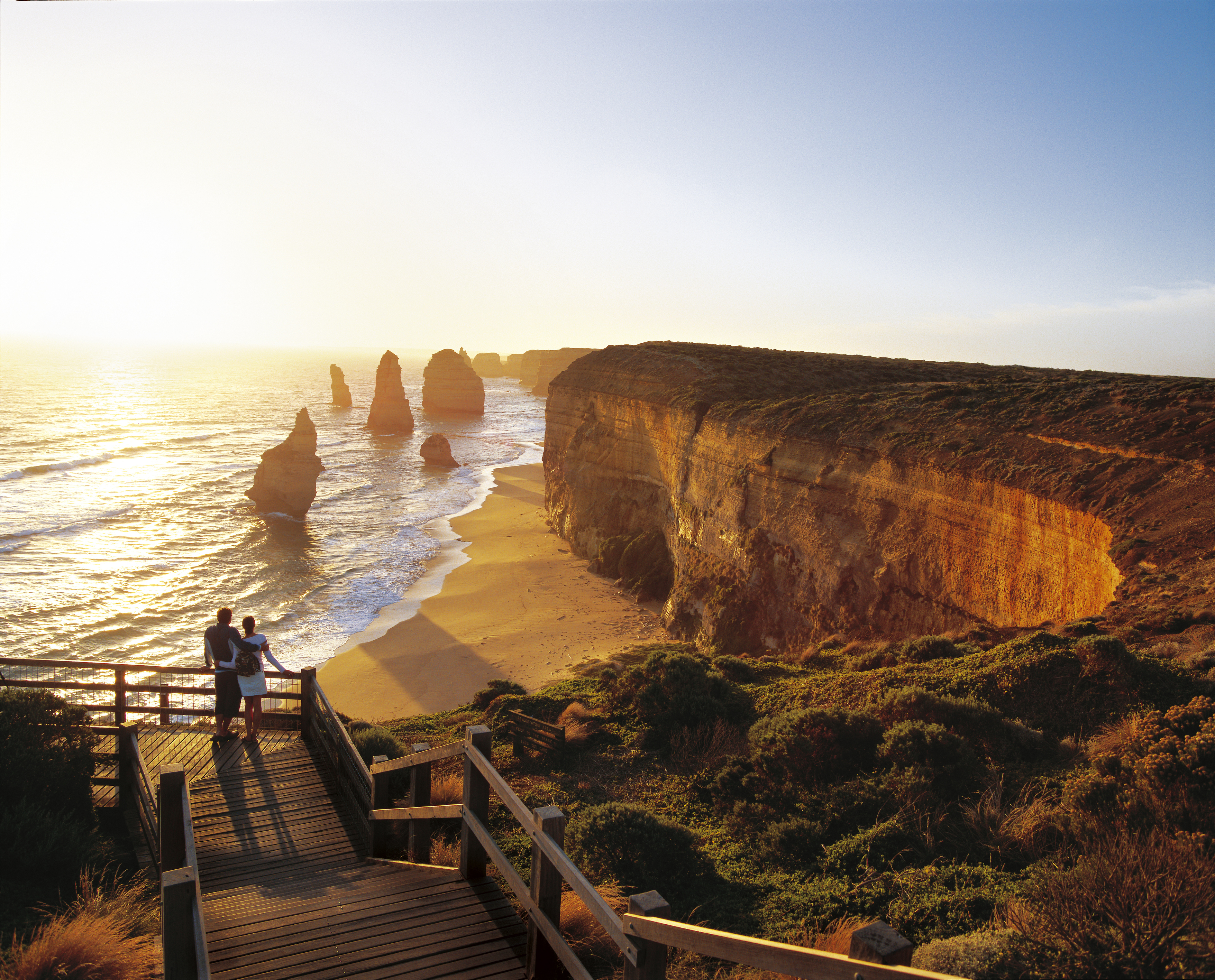 Man and woman admiring the sea at sunset. In the distance are the Twelve Apostles, unique rock formations that rise out of the sea along the Victorian coast.