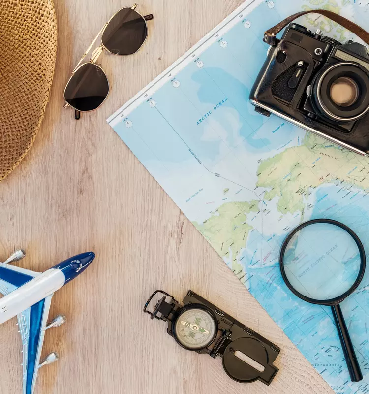 Flat lay of travel essentials including a straw hat, aviator sunglasses, vintage camera, world map, magnifying glass, compass, and a toy airplane on a wooden surface