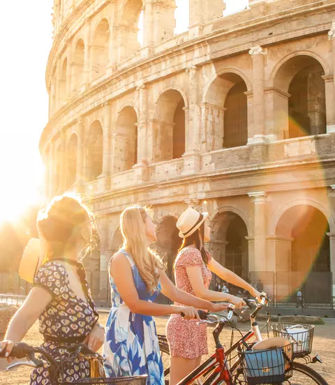 Three happy young women friends tourists with bikes at Colosseum in Rome, Italy at sunrise