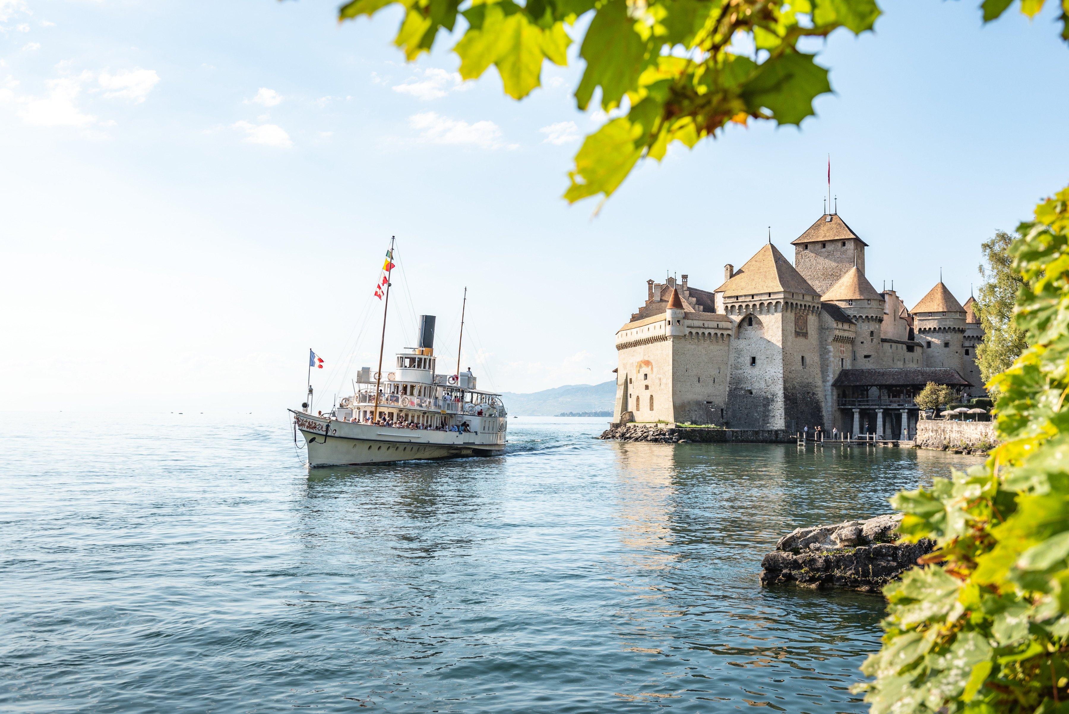 Château de Chillon A vintage steamship sails near the historic Château de Chillon, a medieval castle on the shore of a clear lake, framed by green leaves.