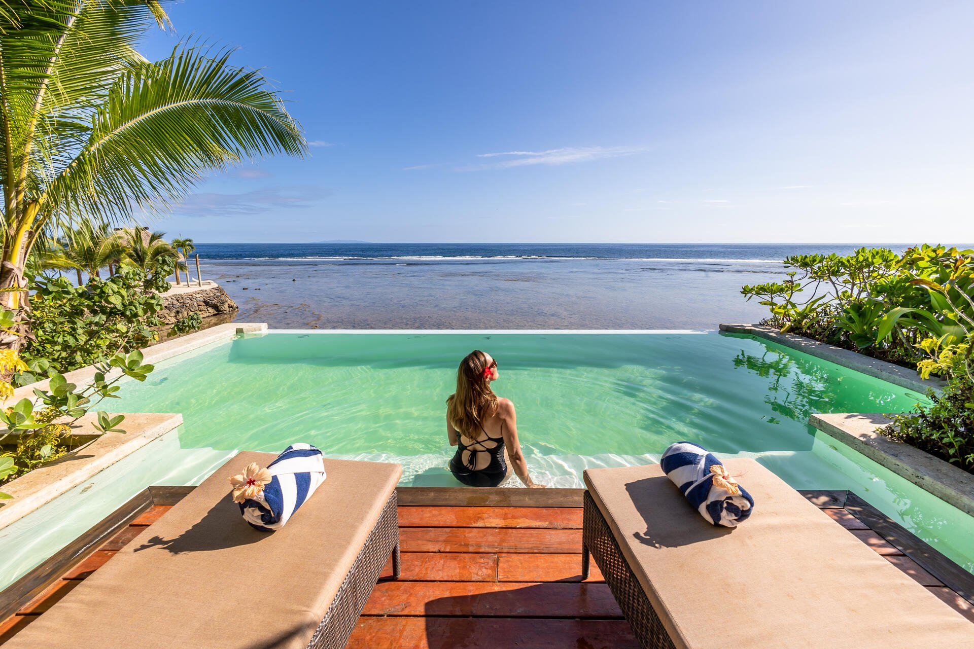 Woman lounging by a beach-side pool, surrounded by palm trees, clear, blue water and sunny skies.