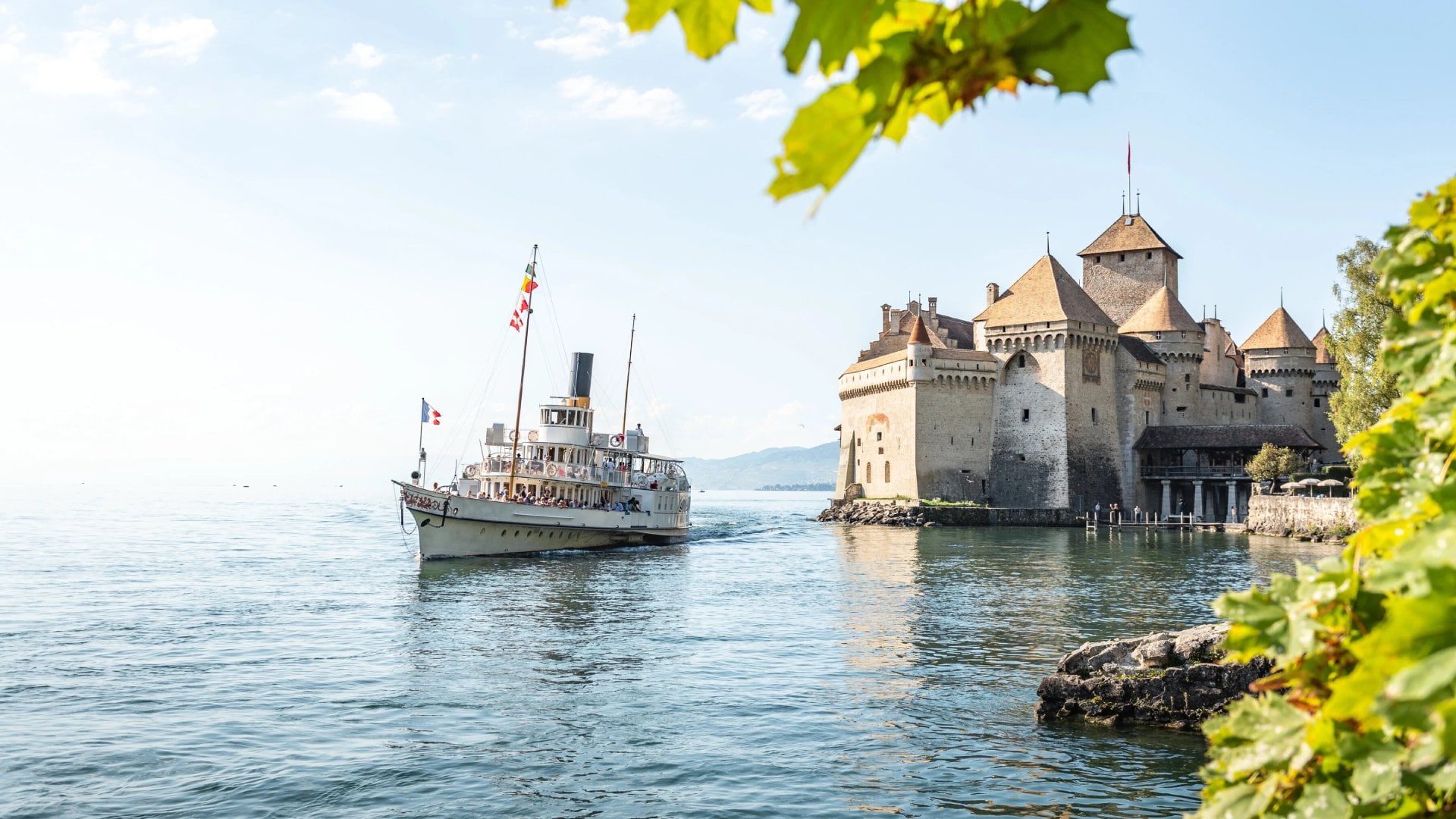 A vintage steamship sails near the historic Château de Chillon, a medieval castle on the shore of a clear lake, framed by green leaves.