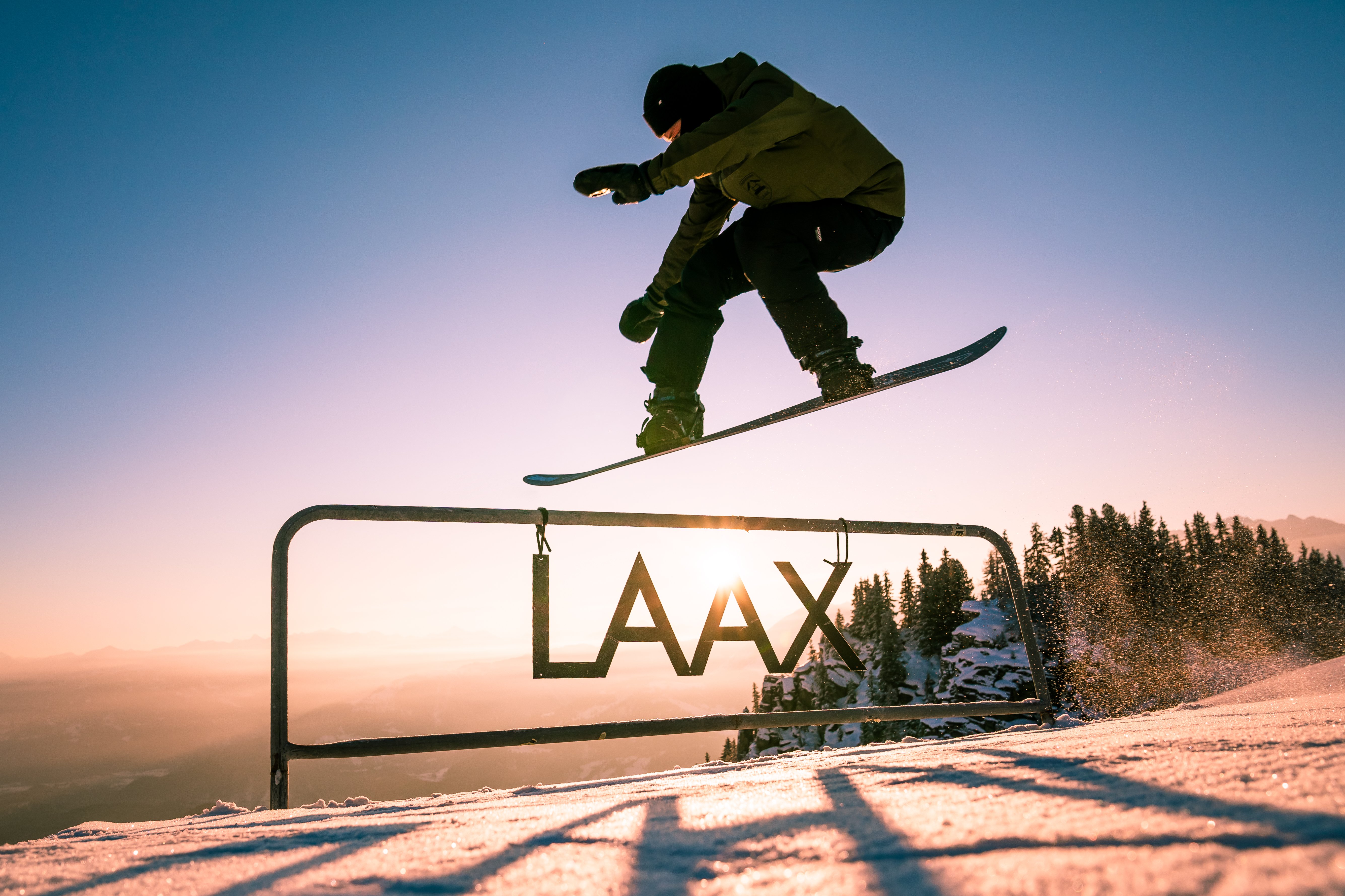 A snowboarder jumping over a sign saying "Laax" at sunset, in Laax, Switzerland.