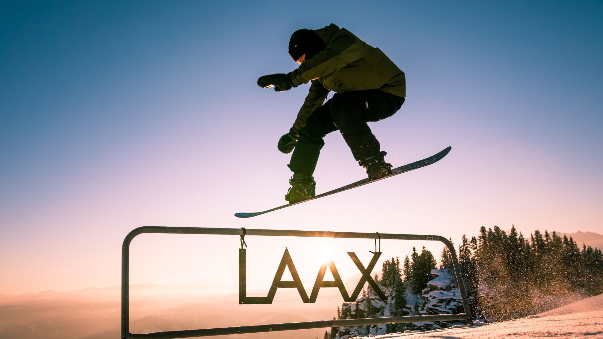 A snowboarder jumping over a sign saying "Laax" at sunset, in Laax, Switzerland.