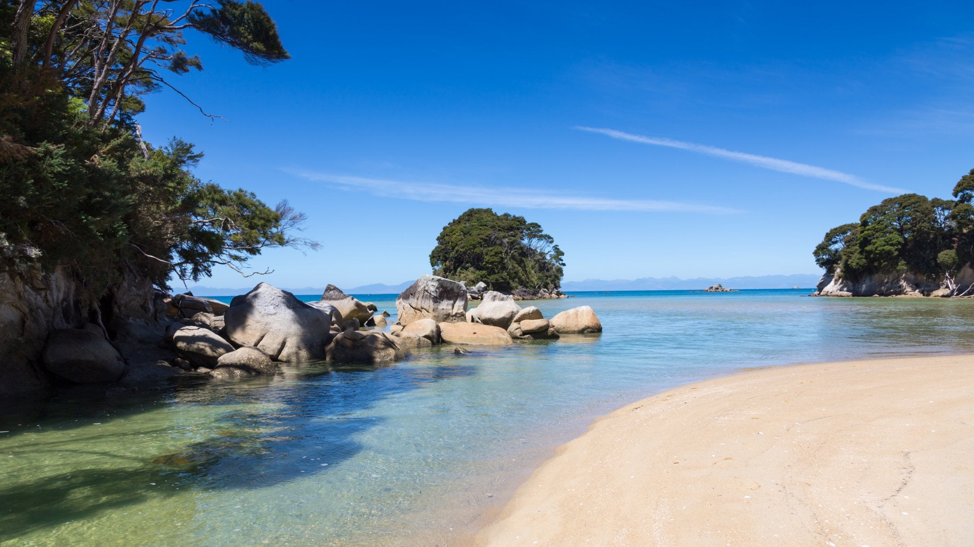 New Zealand Abel Tasman National Park on South Island New Zealand on the beach on day time. The water is calm and clear. There is big rocks into the water and few islands. The sky is clear blue.