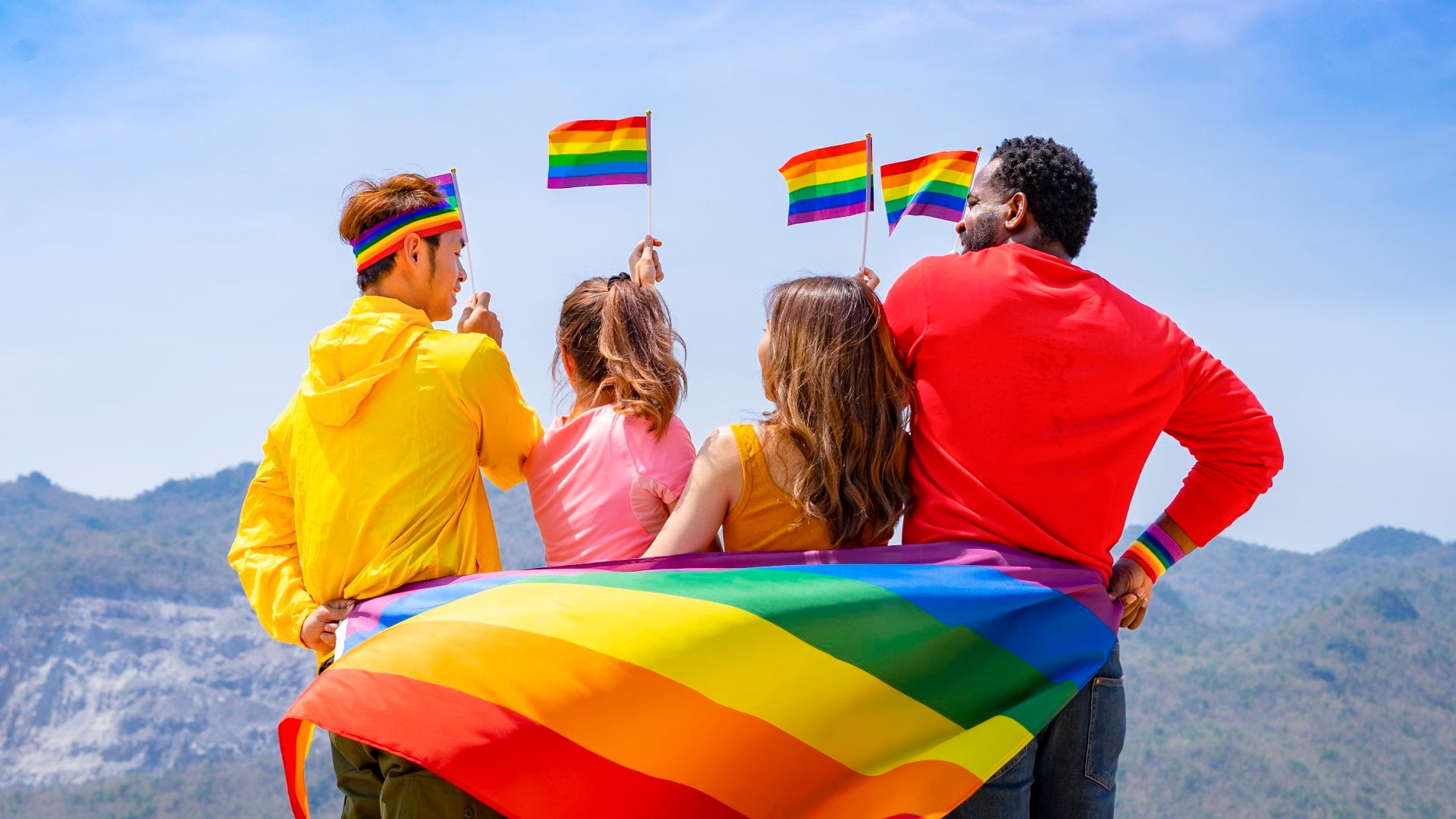 Group of diverse lgbtq friends wearing colorful clothes, standing side by side, smiling and holding lgbt flags waving in the air