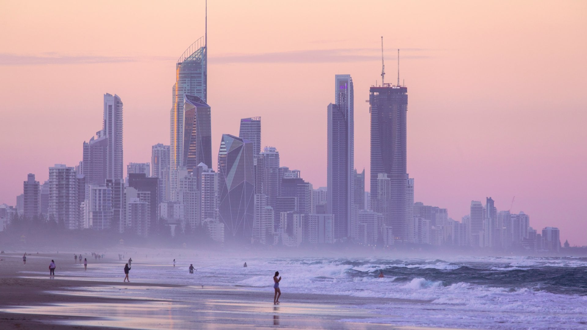 People walk along the beach at twilight against a backdrop of the Gold Coast skyscrapers in Australia.