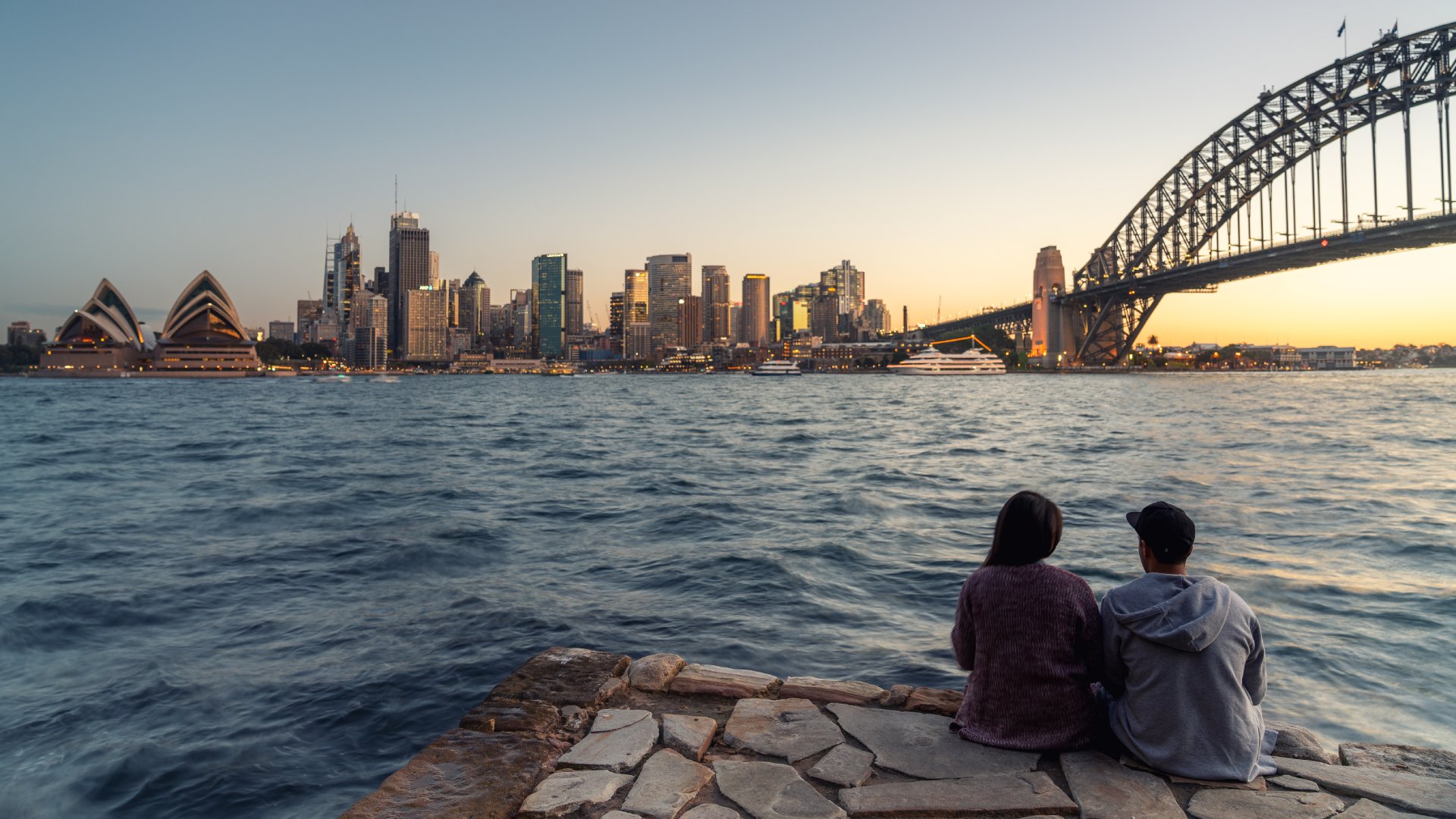 couple sitting on edge of harbour looking at bridge and skyline