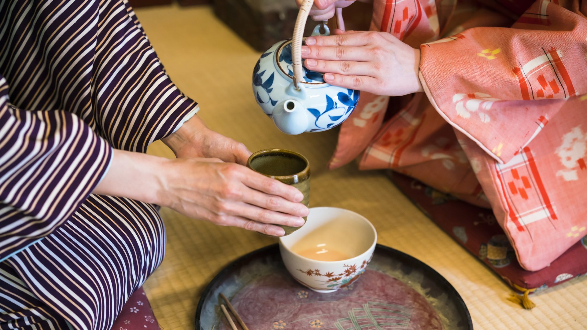 detail of two women in traditional kimono, kneeling on tatami preparing, pouring a cup of tea which is inthe hands of one woman. They are in traditional Japanese old house on tatami. This is in Toei studios in Kyoto with old buildings from Samurai times.
