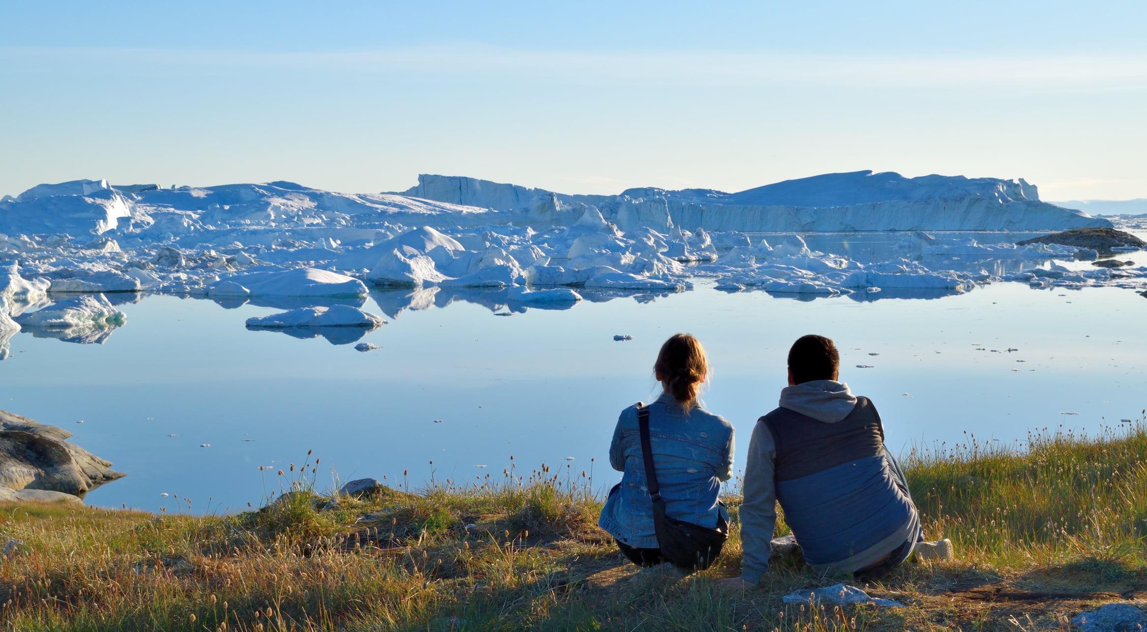 A man and woman sitting together on a field of grass in the foreground, with a frigid looking lake in front of them, leading to a vast ice sheet in  in Ilulissat, Greenland.