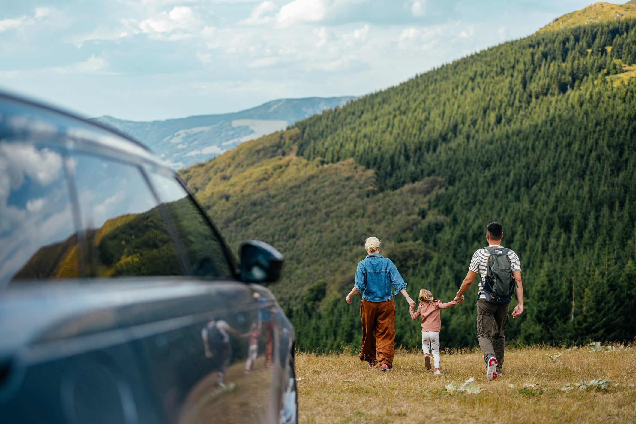 A family holding hands starting a forest hike with their car in the foreground.