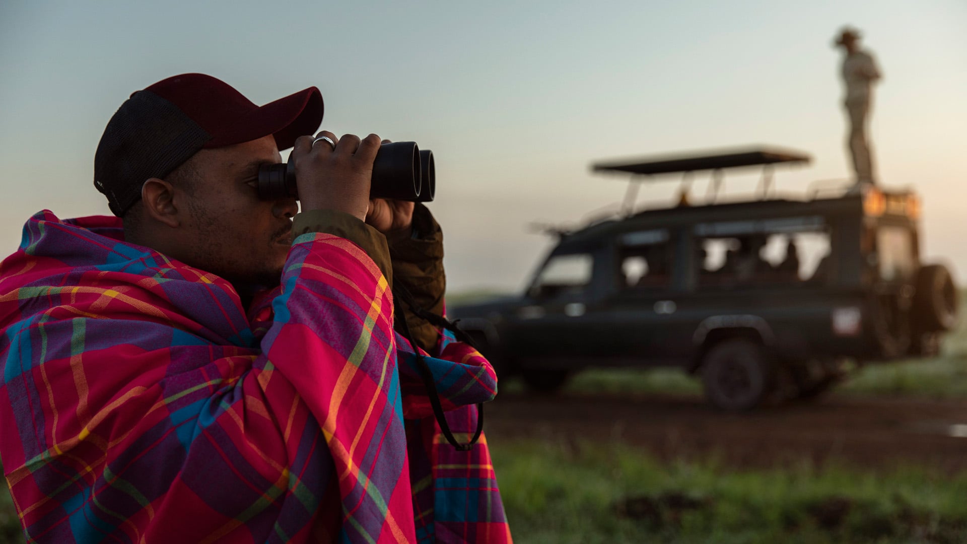 A Maasai safari tracker looks through binoculars while someone stands on a land cruiser in the background in Kenya.
