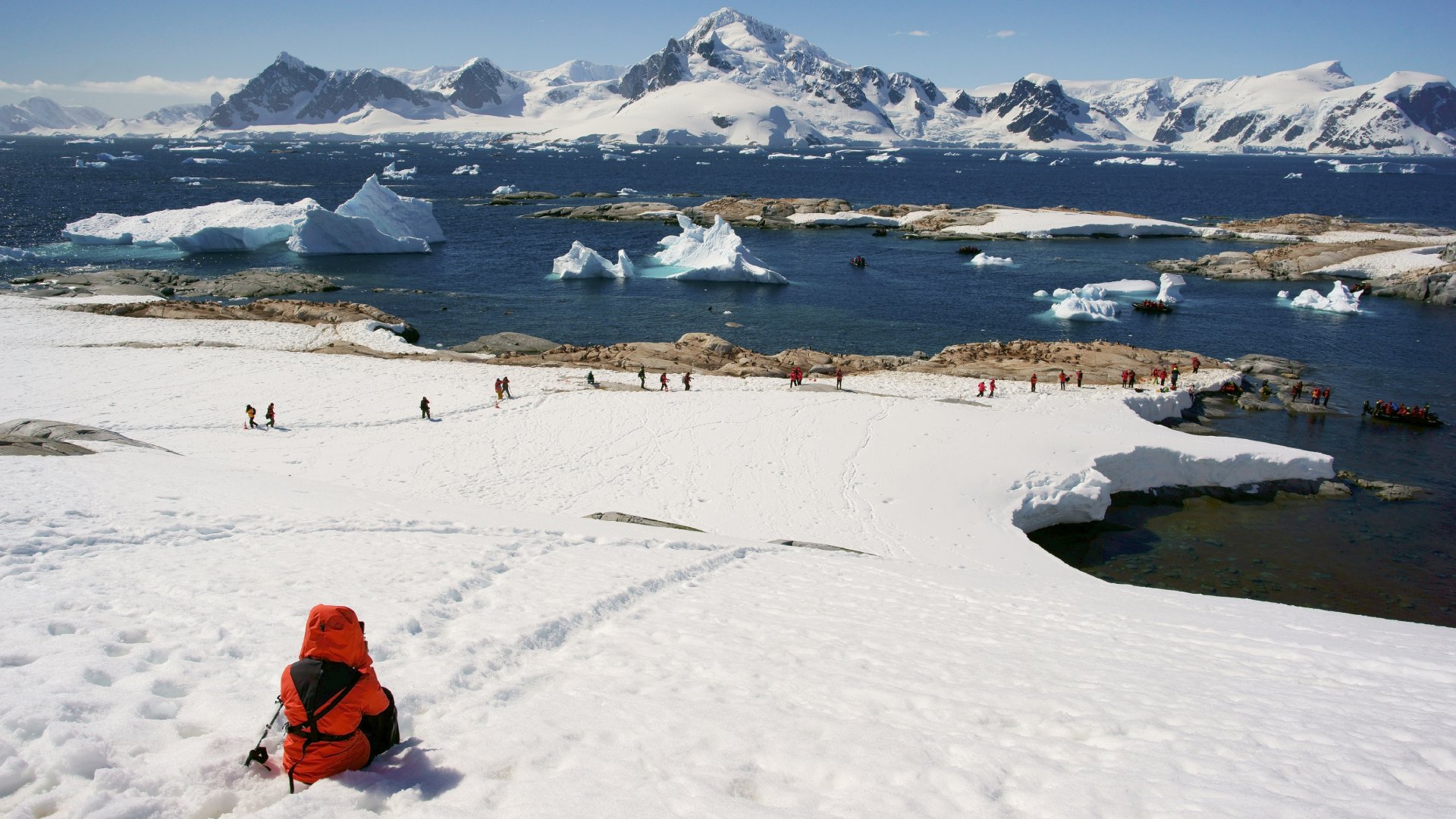 A person in a bright orange jacket sits on snowy terrain, overlooking a glacier and a group walking near the water.