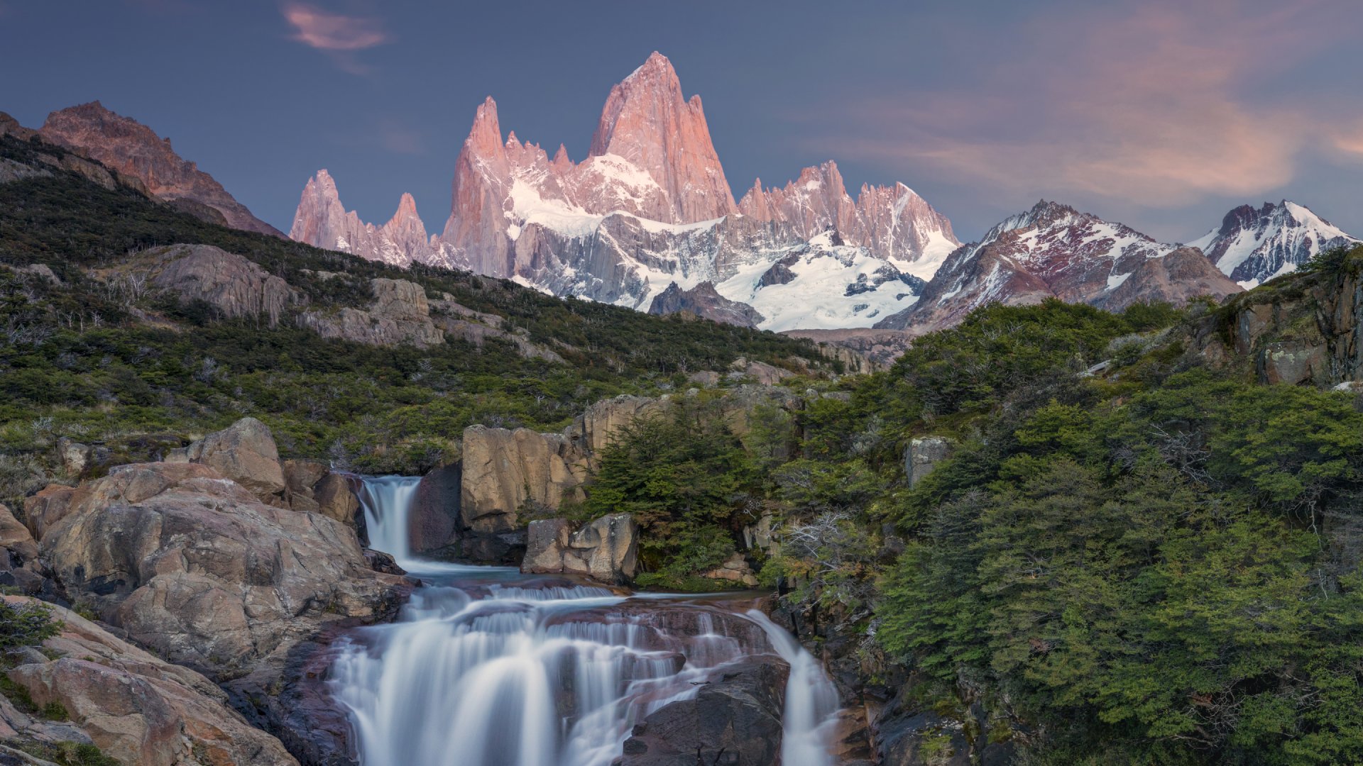 A serene landscape featuring a waterfall flowing through rocks, with majestic mountains and a colorful sky in the background.