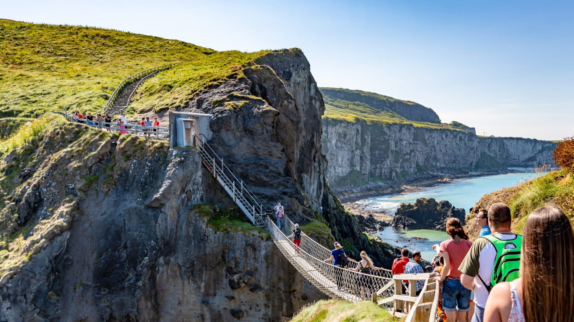 A sunny coastal scene showing a suspended bridge with people walking towards scenic cliffs and the ocean in the background.