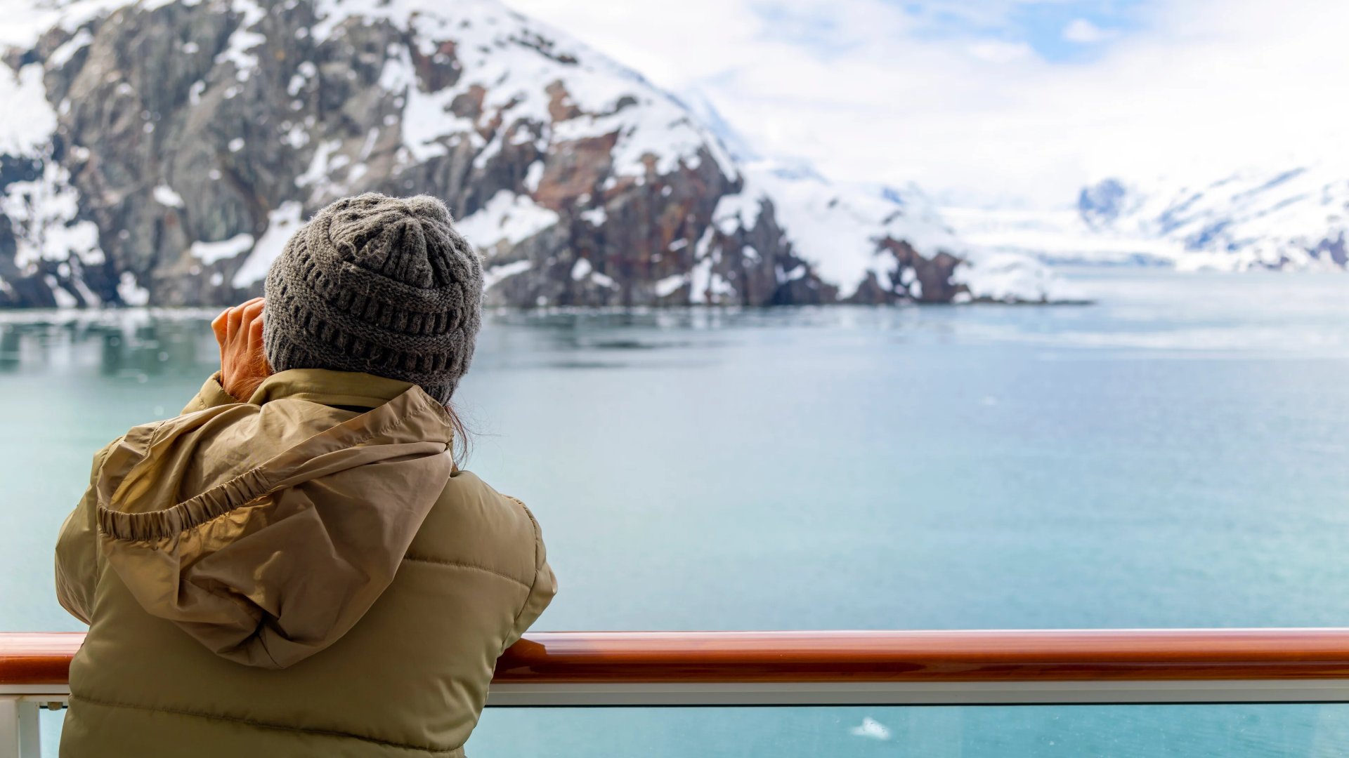 A young woman with binoculars views the snow covered mountains and glaciers from a cruise ship balcony at Glacier Bay National Park and Reserve, Alaska USA.