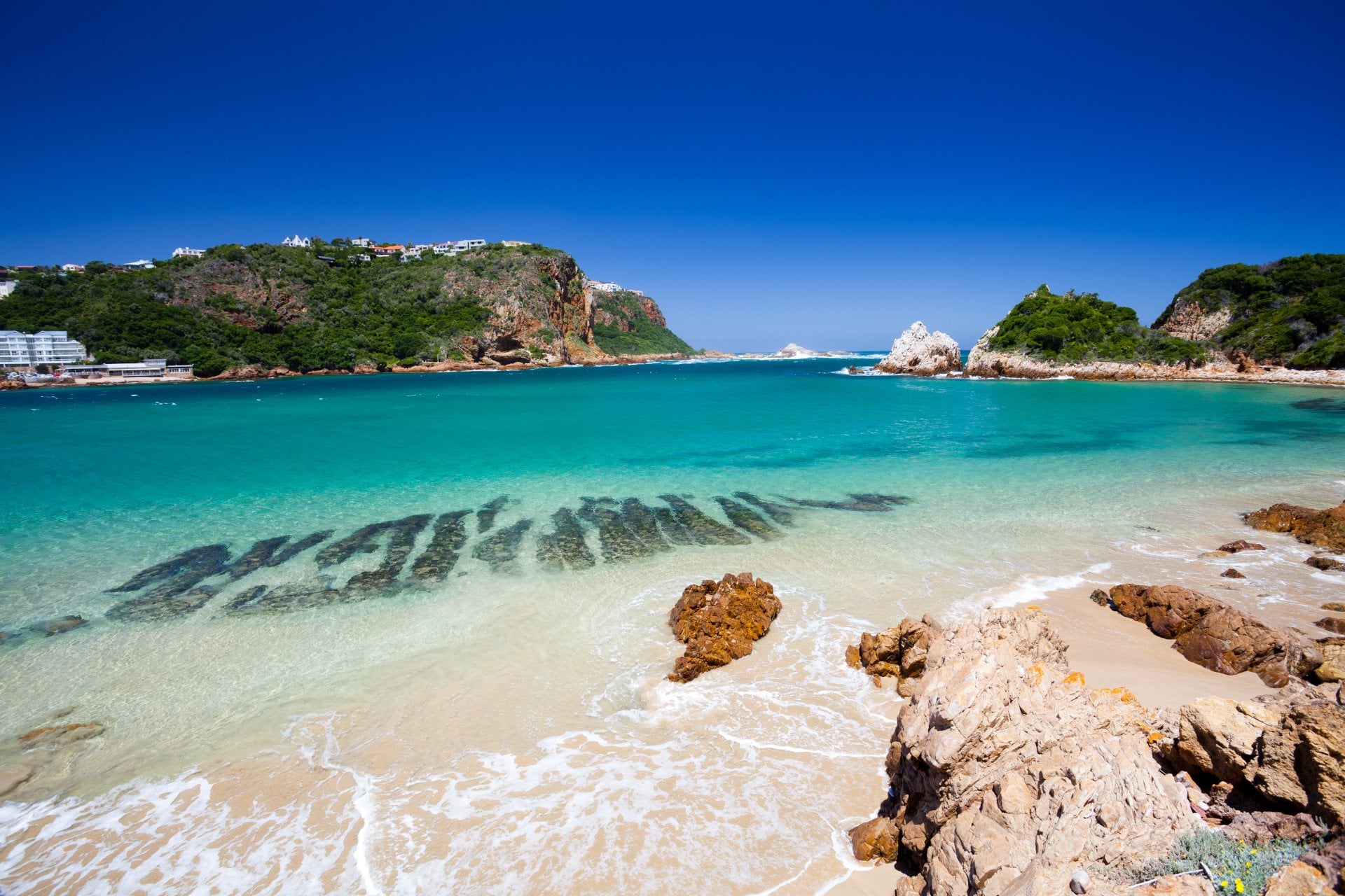 A beach in Knysna, Western Cape, South Africa. The beach is surrounded by lush green hills and has white sand and clear blue water.