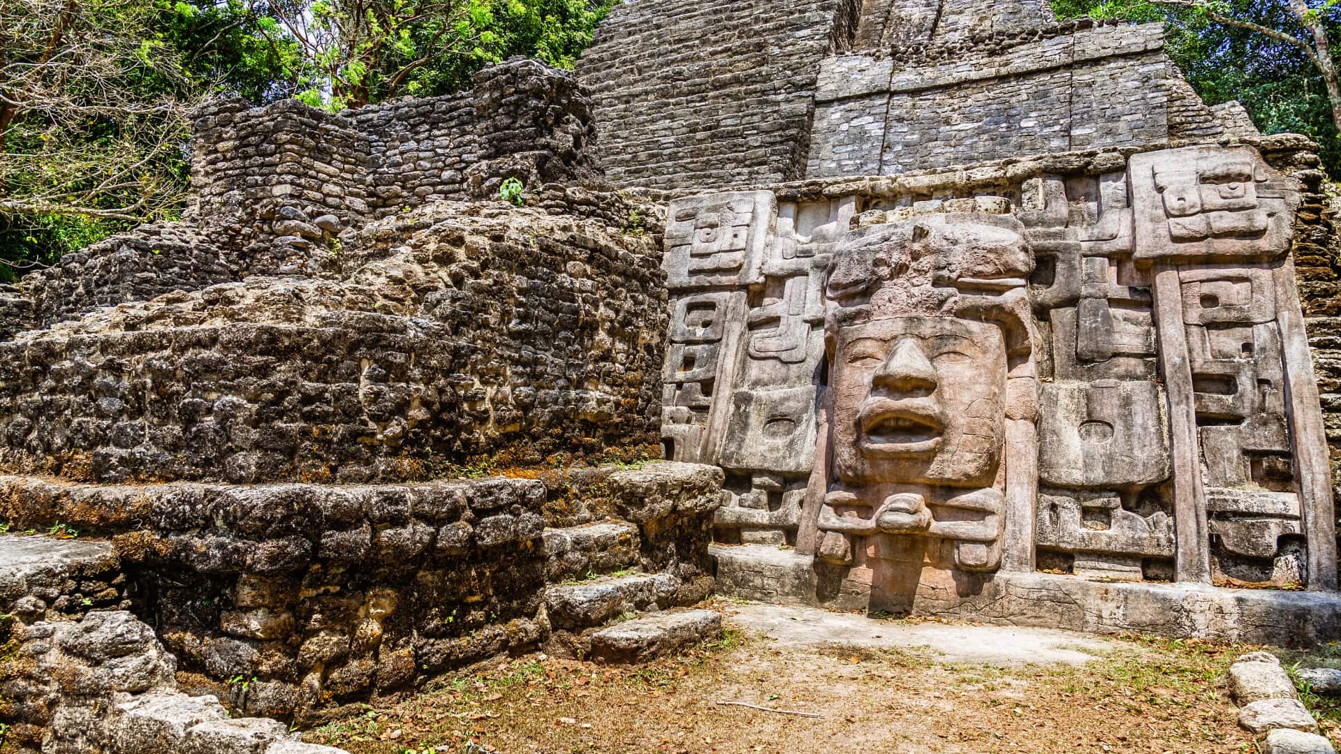 Ancient stone ruins with a carved face, surrounded by greenery and blue sky.