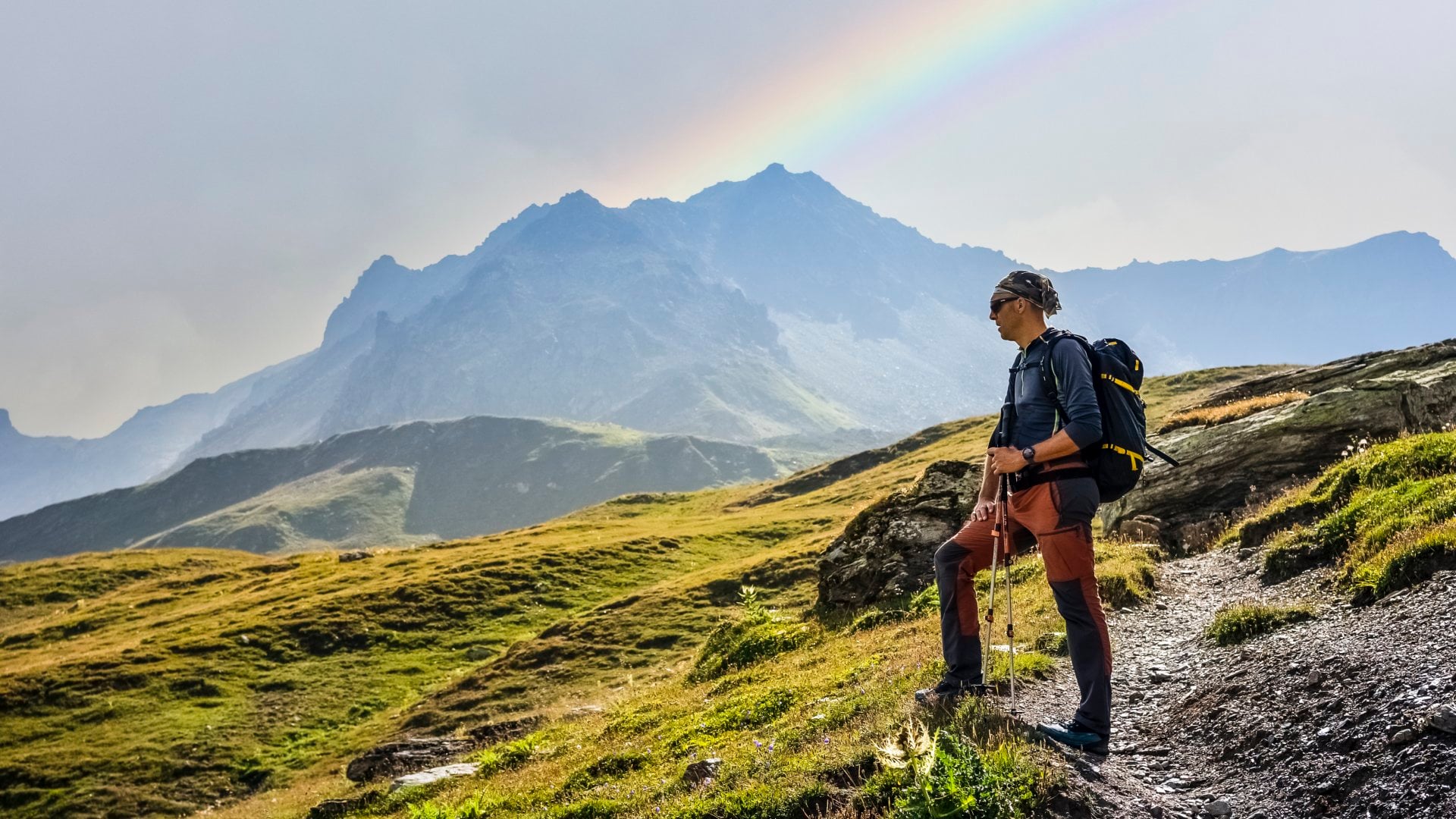 Hiker standing on a rocky trail, gazing at mountains and a rainbow in the sky.