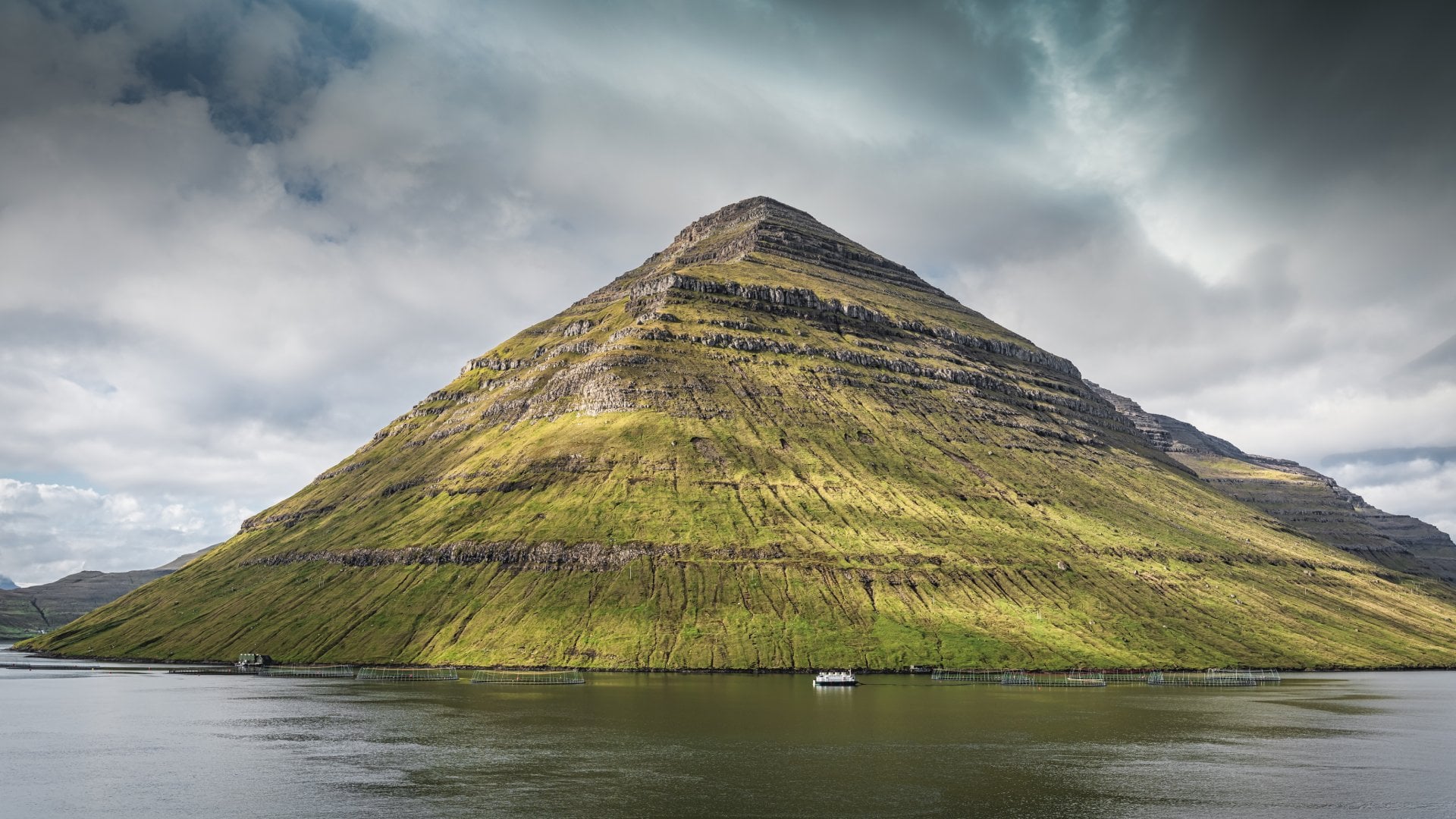 Majestic Pyramid Shaped tall North Atlantic Ocean Fjord Mountain of Kunoy Island under moody cloudscape with sunlight on the mountain
