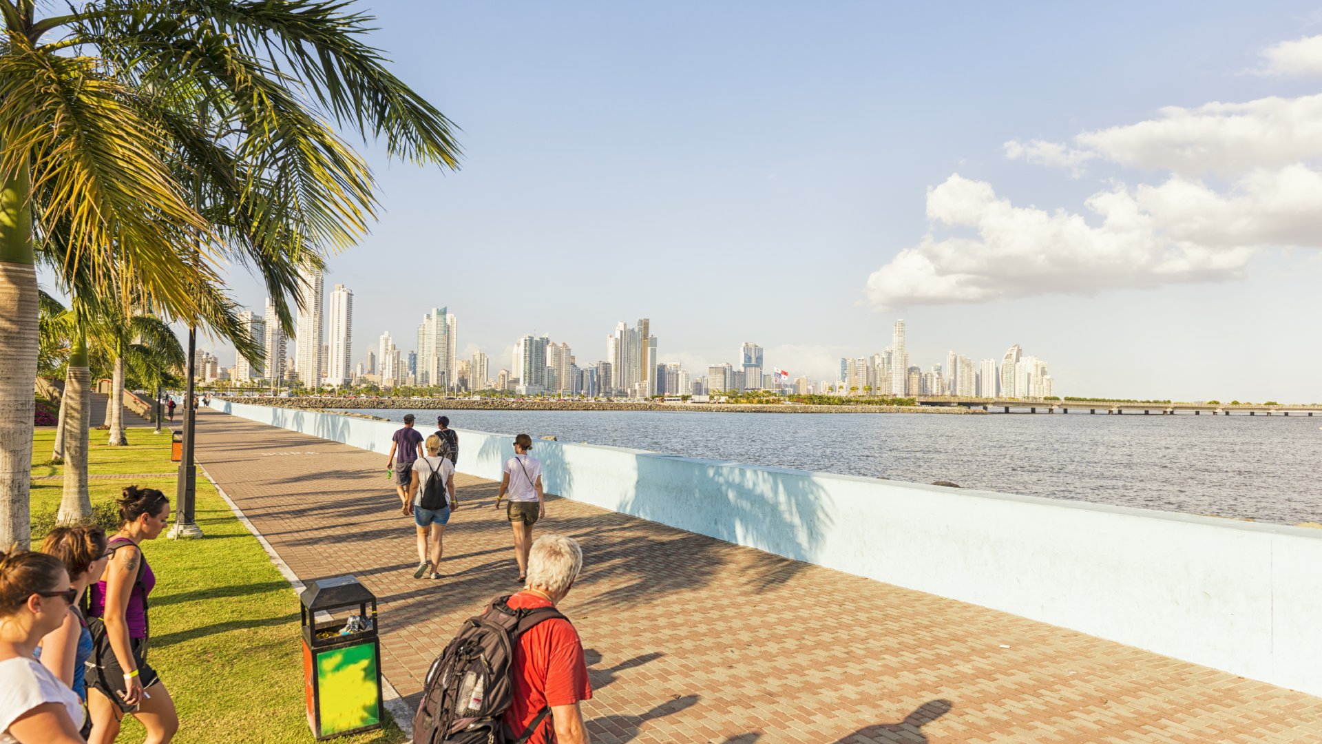People walking along a waterfront promenade with a skyline and palm trees in the background under a clear sky.