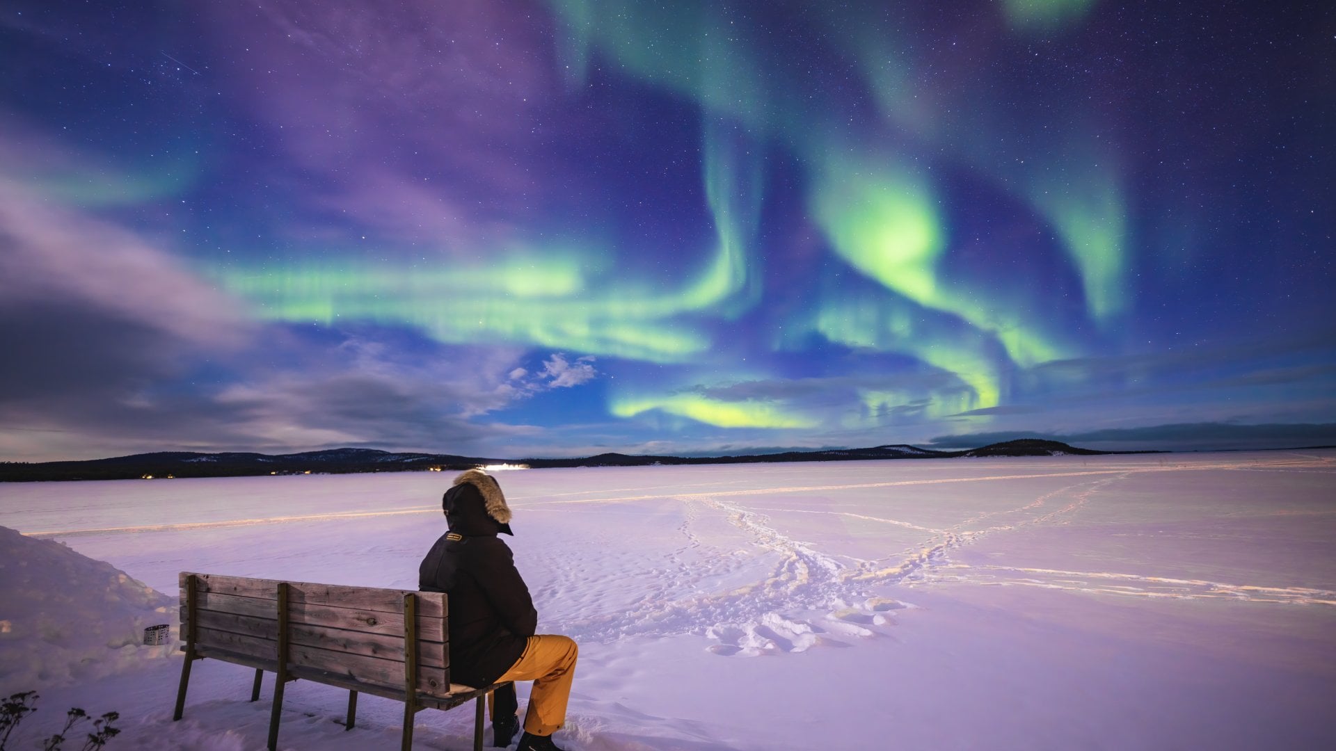 Person sitting on a bench, gazing at vibrant northern lights over a snowy landscape.