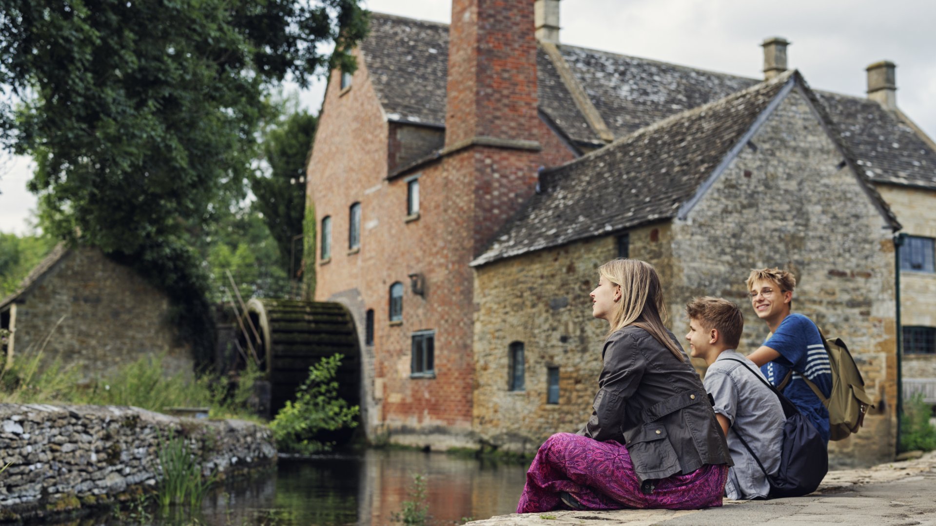 Three children sit by a river, enjoying the view of a rustic building and a waterwheel in a peaceful countryside setting.