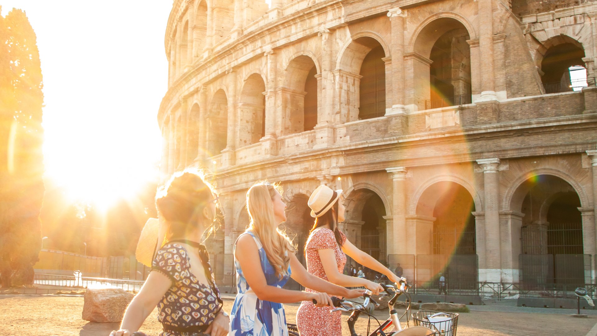 Three happy young women friends tourists with bikes at Colosseum in Rome, Italy at sunrise