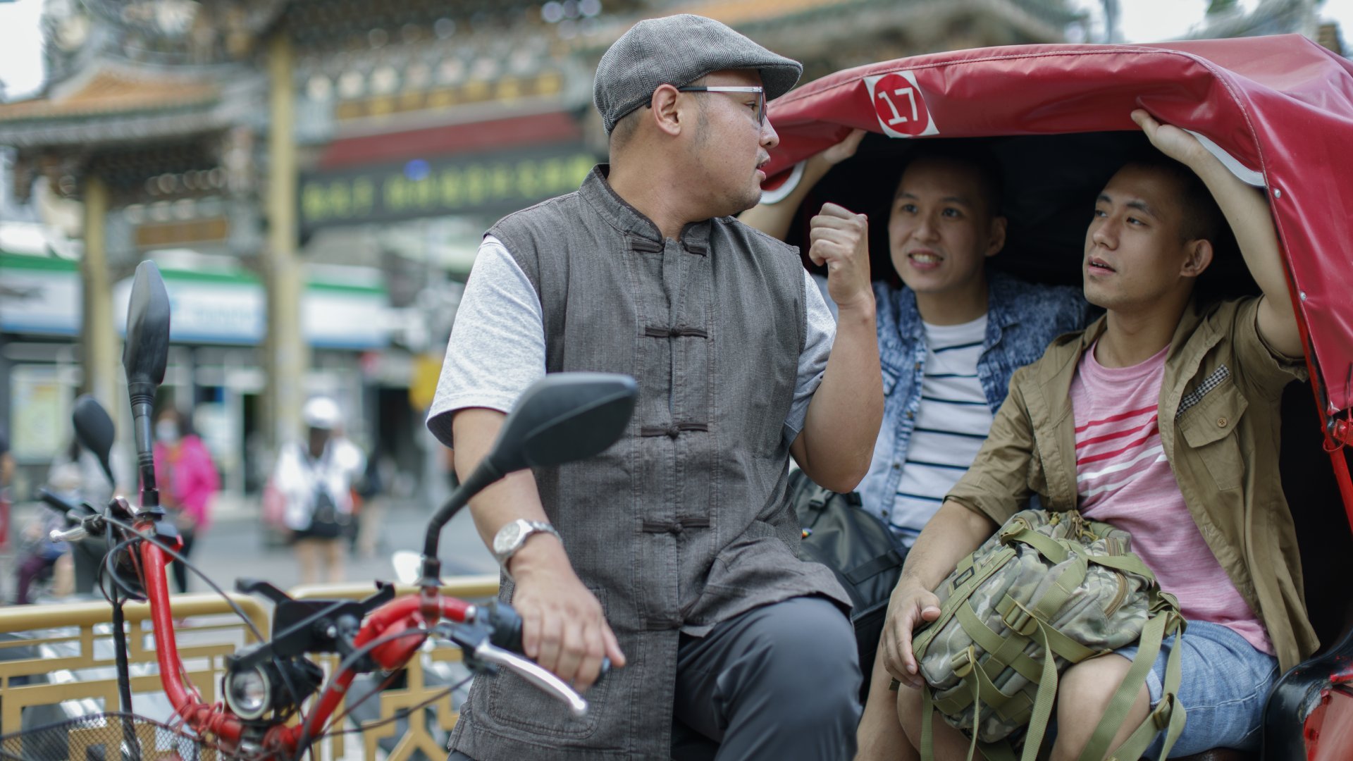 Three people interact in a rickshaw; a driver wearing a cap chatters with two passengers in a busy street setting.