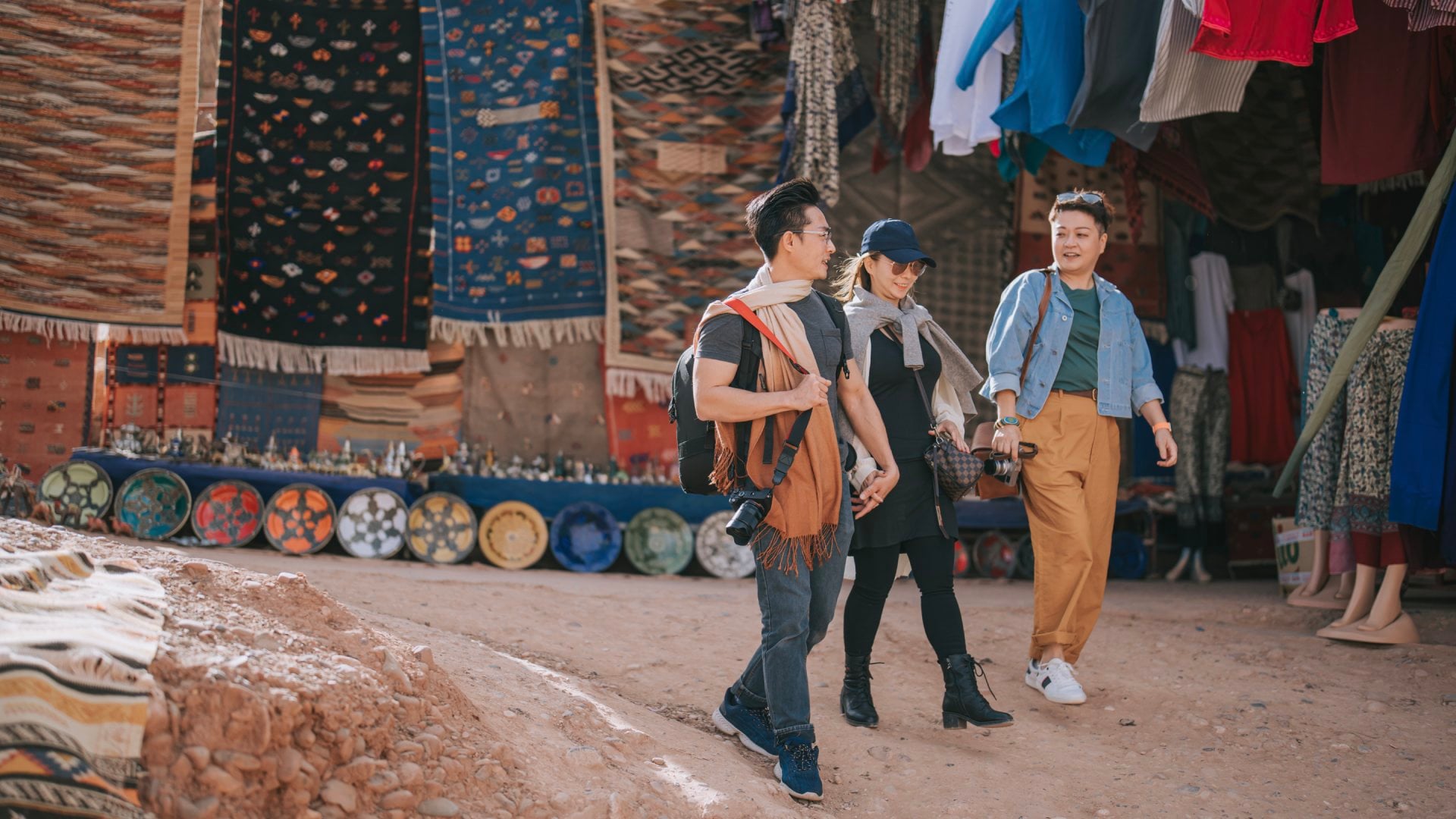 Three people walk through a vibrant market, surrounded by colorful textiles and handmade pottery.