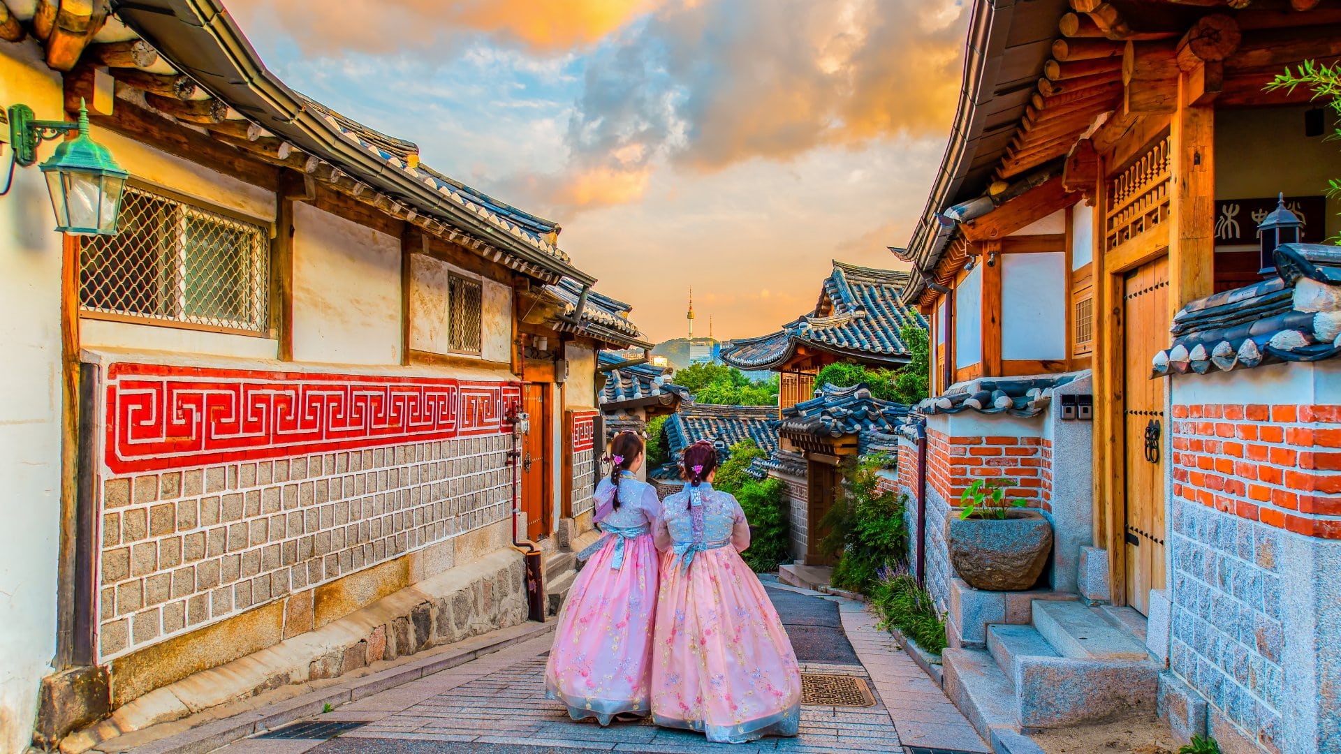Two girls in traditional attire stroll through a picturesque alley lined with wooden houses and colorful sky at sunset.