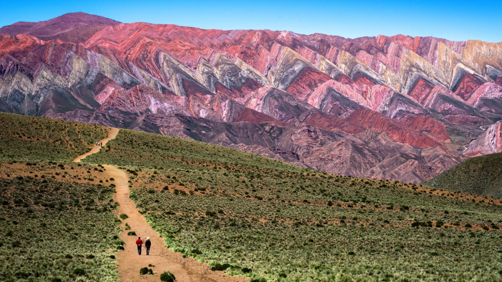 Two hikers walk along a dirt path with colorful striped mountains in the background under a clear blue sky.