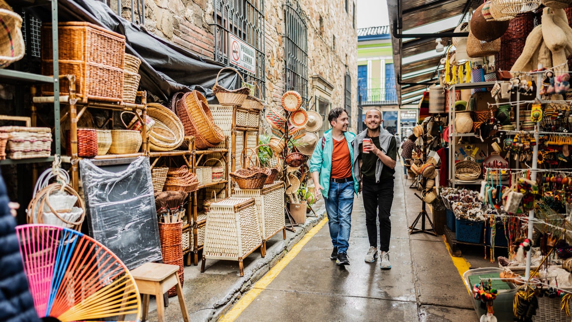 Two men stroll through a vibrant market alley filled with colorful handmade crafts and woven baskets.