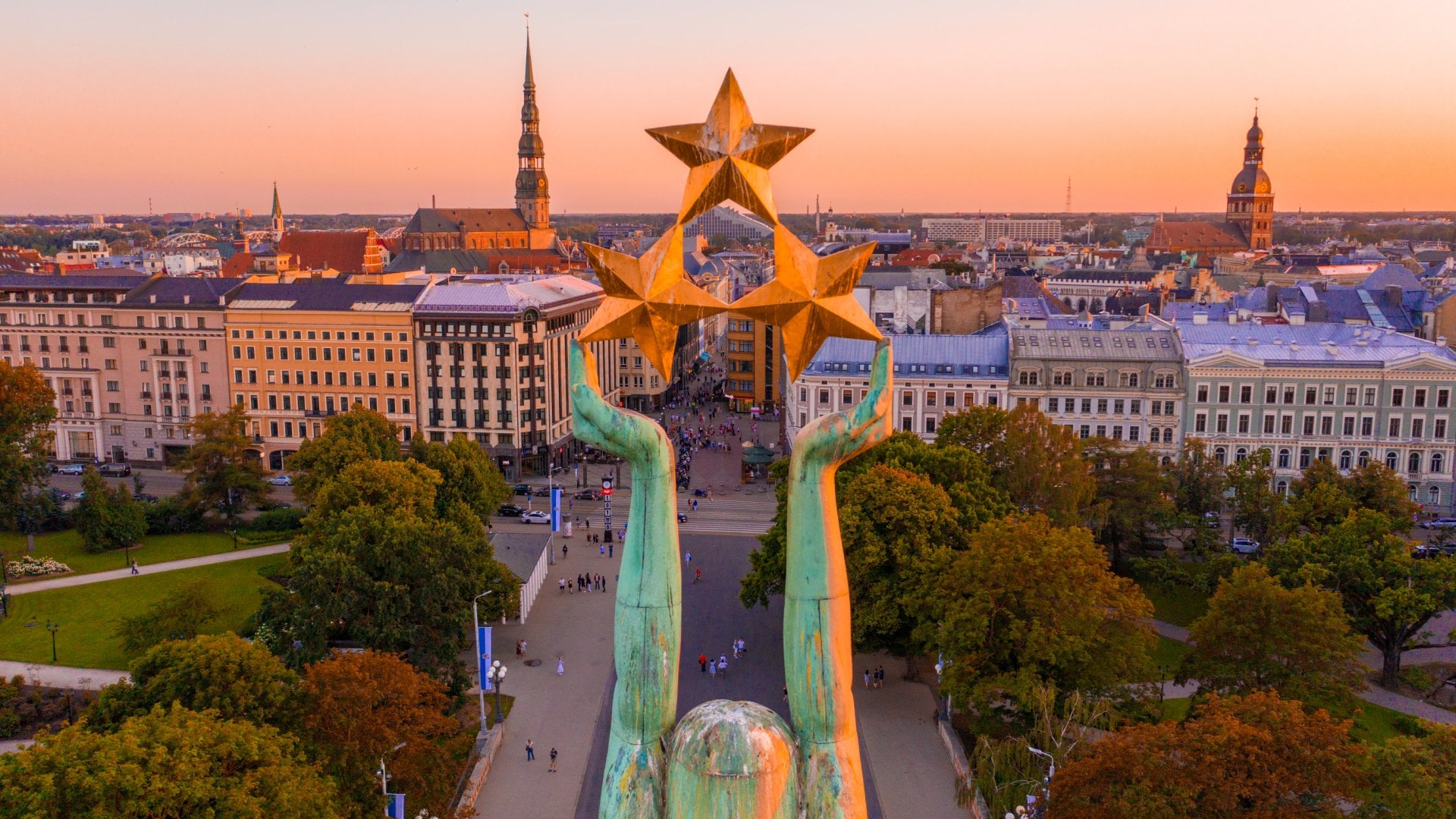 View of a city at sunset, featuring a monumental statue with raised hands holding three stars, surrounded by trees and buildings.