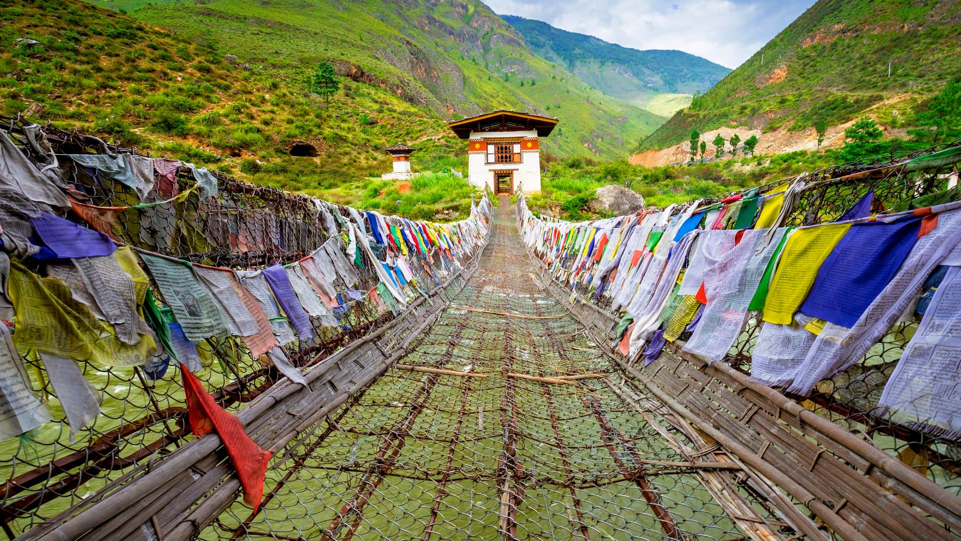 View straight on of Iron Chain Bridge of Tamchog Lhakhang Monastery with prayer flags lining the bridge, mountains and cloudy sky in the background