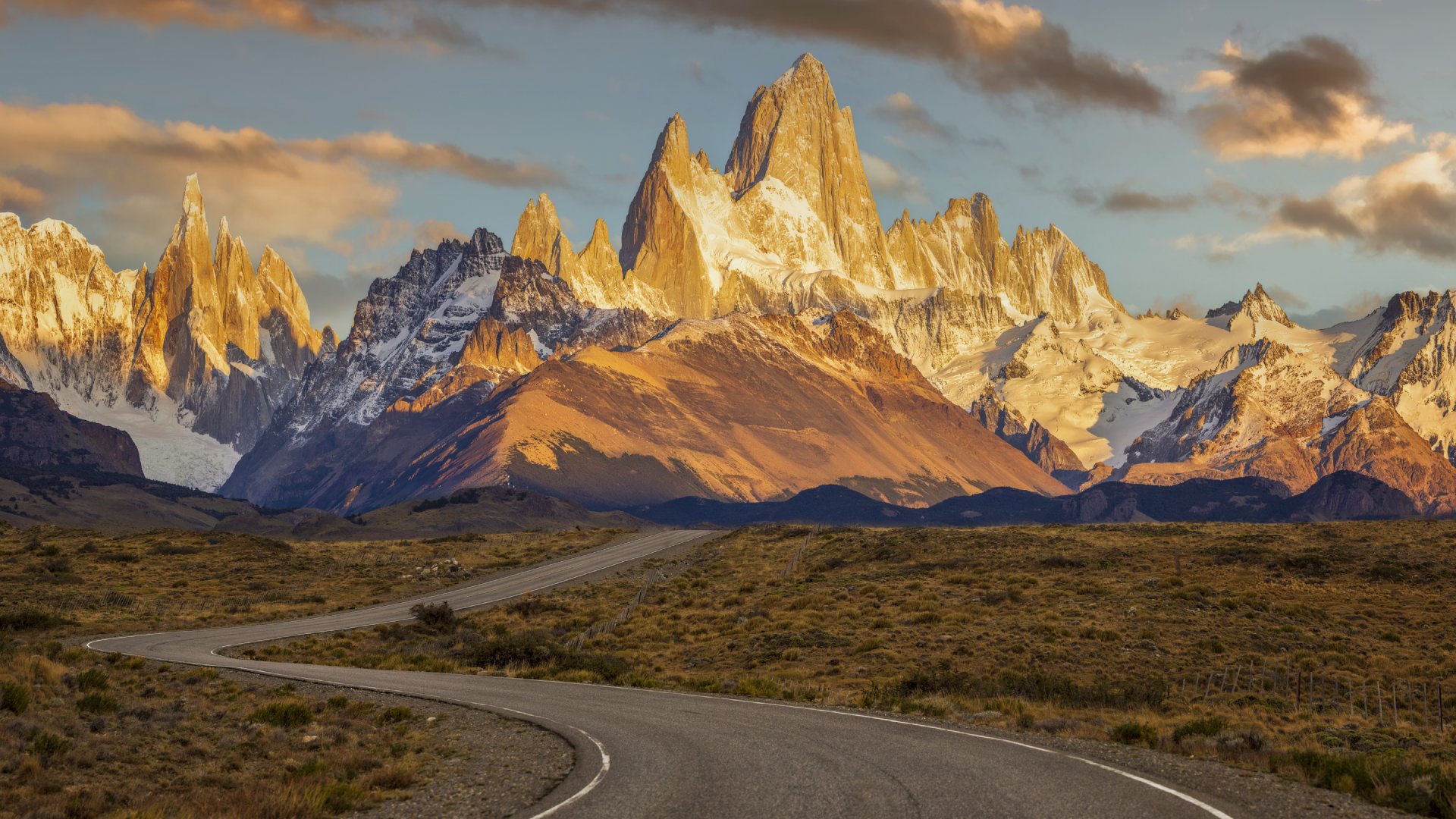 Winding road leads toward dramatic, snow-capped mountains at sunset, with warm, glowing light illuminating the peaks.