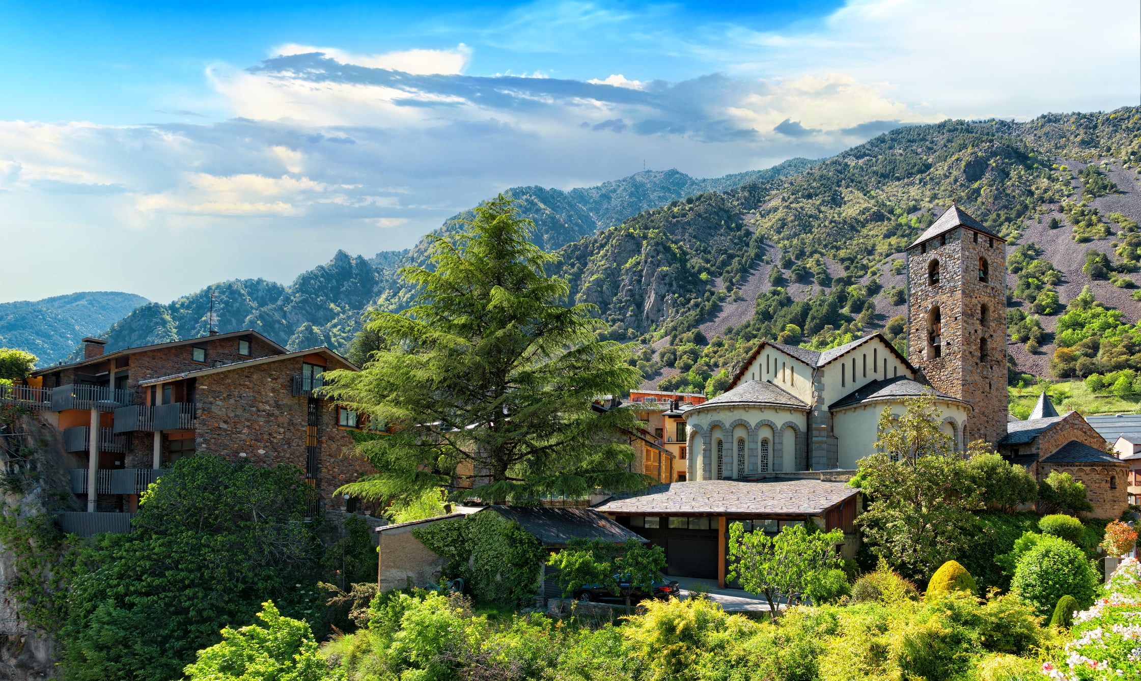 Bright and sunny day over looking lush tree-tops, a mountain range, and a gothic-style building in La Vella, Andorra.