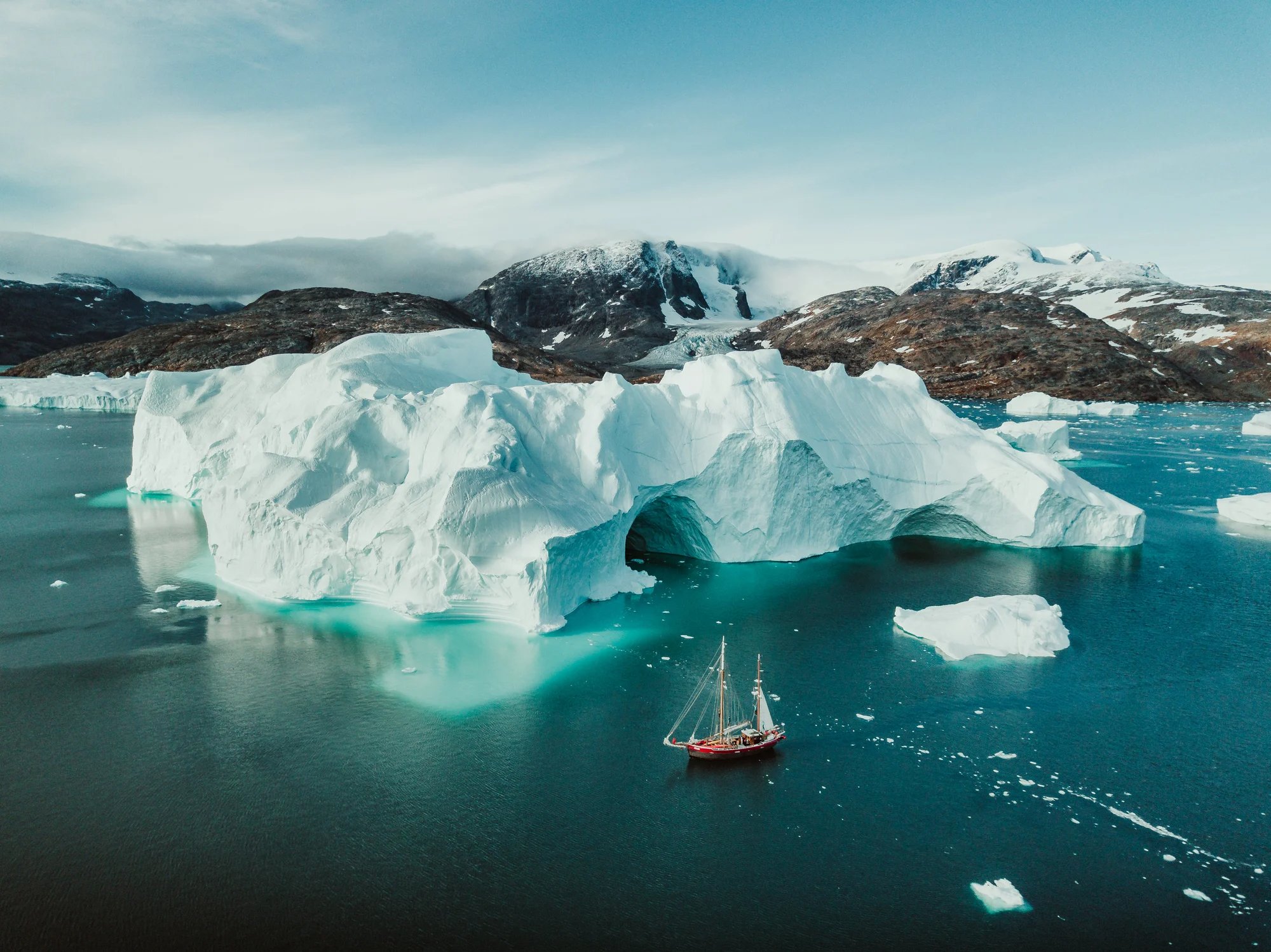 An aerial view of a sailboat passing alongside icebergs in the cold Arctic waters of Greenland.