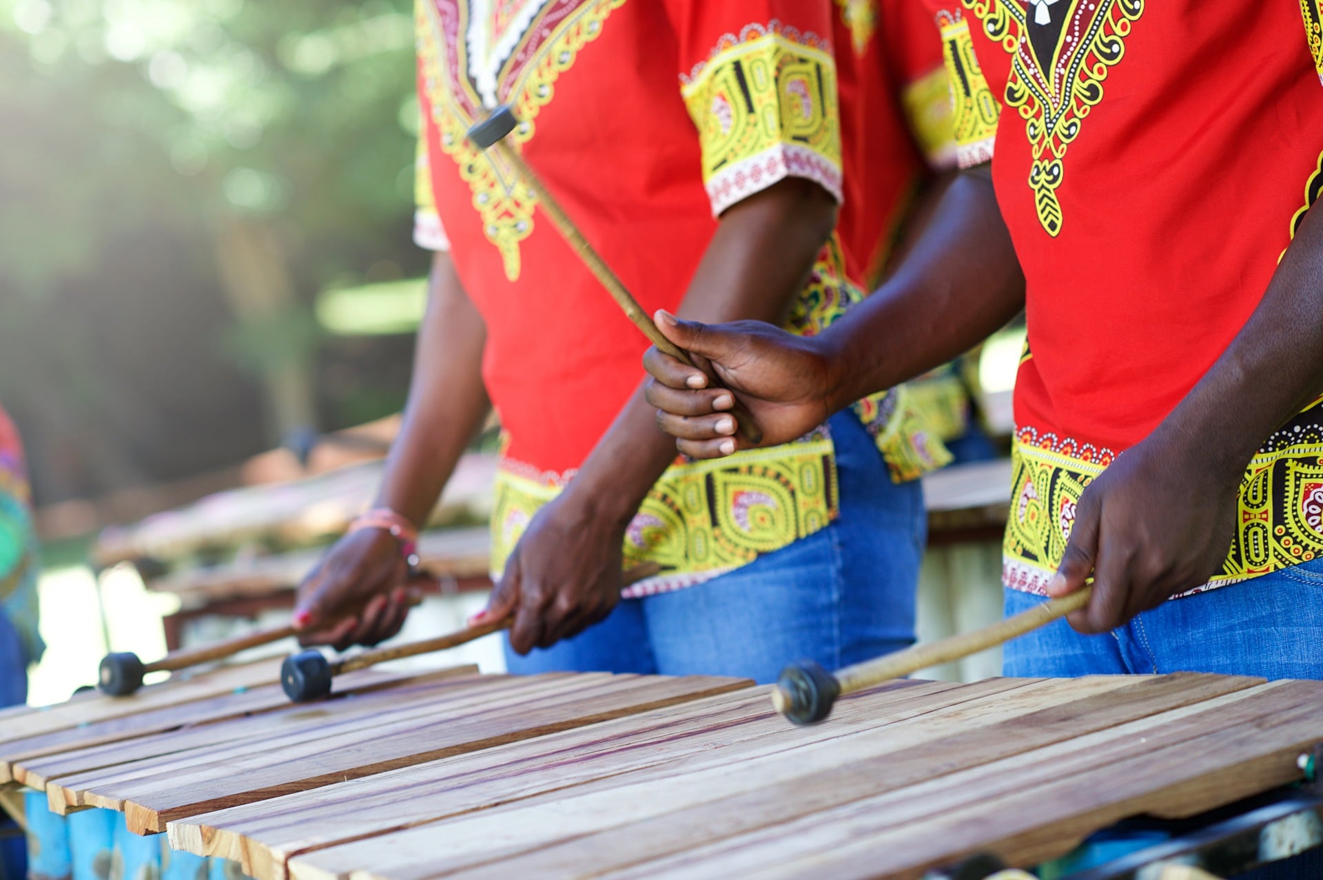 Three musicians in colorful shirts play a wooden xylophone outdoors, holding mallets and striking the bars.
