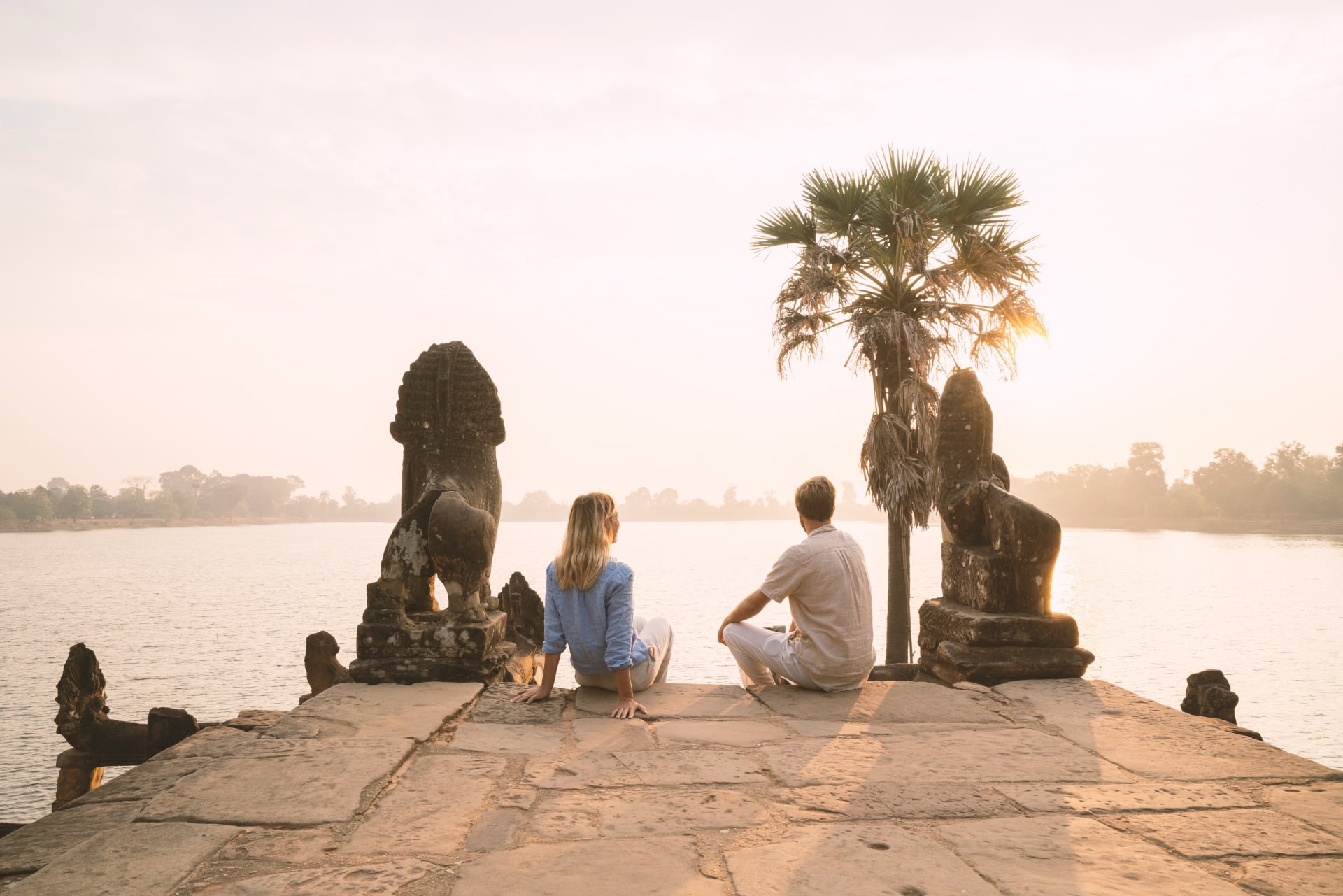 A couple sits on a stone platform, overlooking a calm river at sunrise, flanked by lion statues and a palm tree.