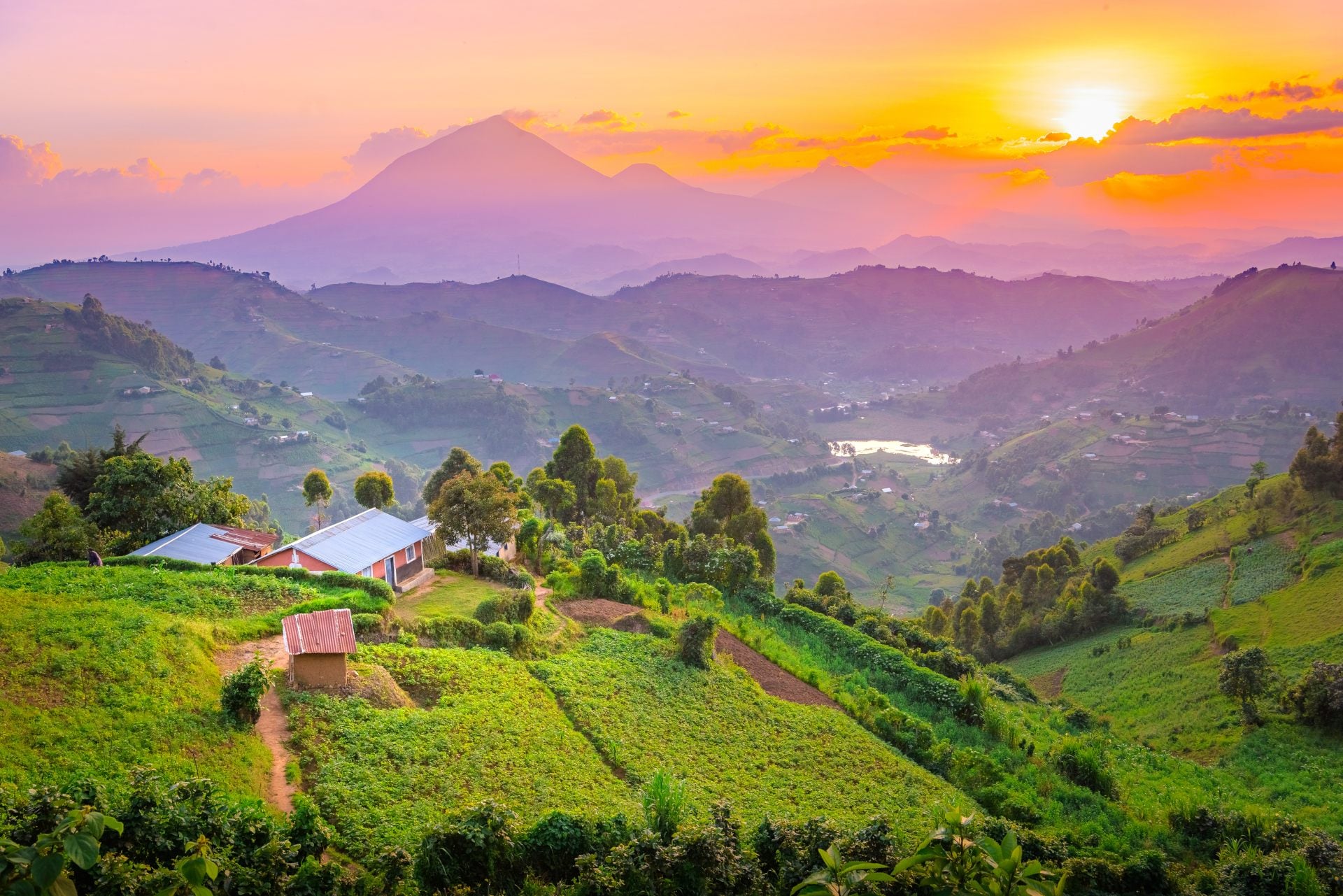 Sunrise over rolling hills with lush greenery and a distant mountain, featuring a small cluster of houses.