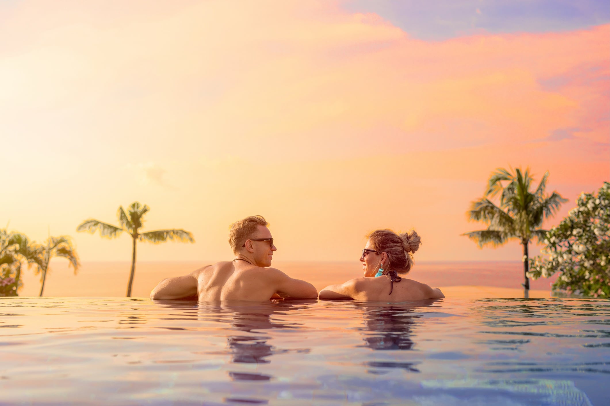 A couple relaxes in an infinity pool, watching a colorful sunset with palm trees in the background.