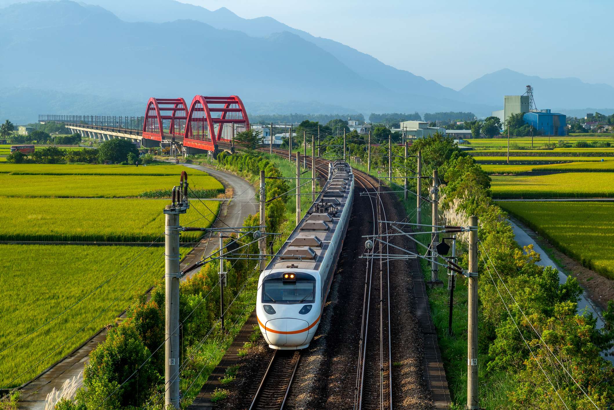 A train travels through a green landscape with rice fields and a red bridge in the background, under a clear blue sky.