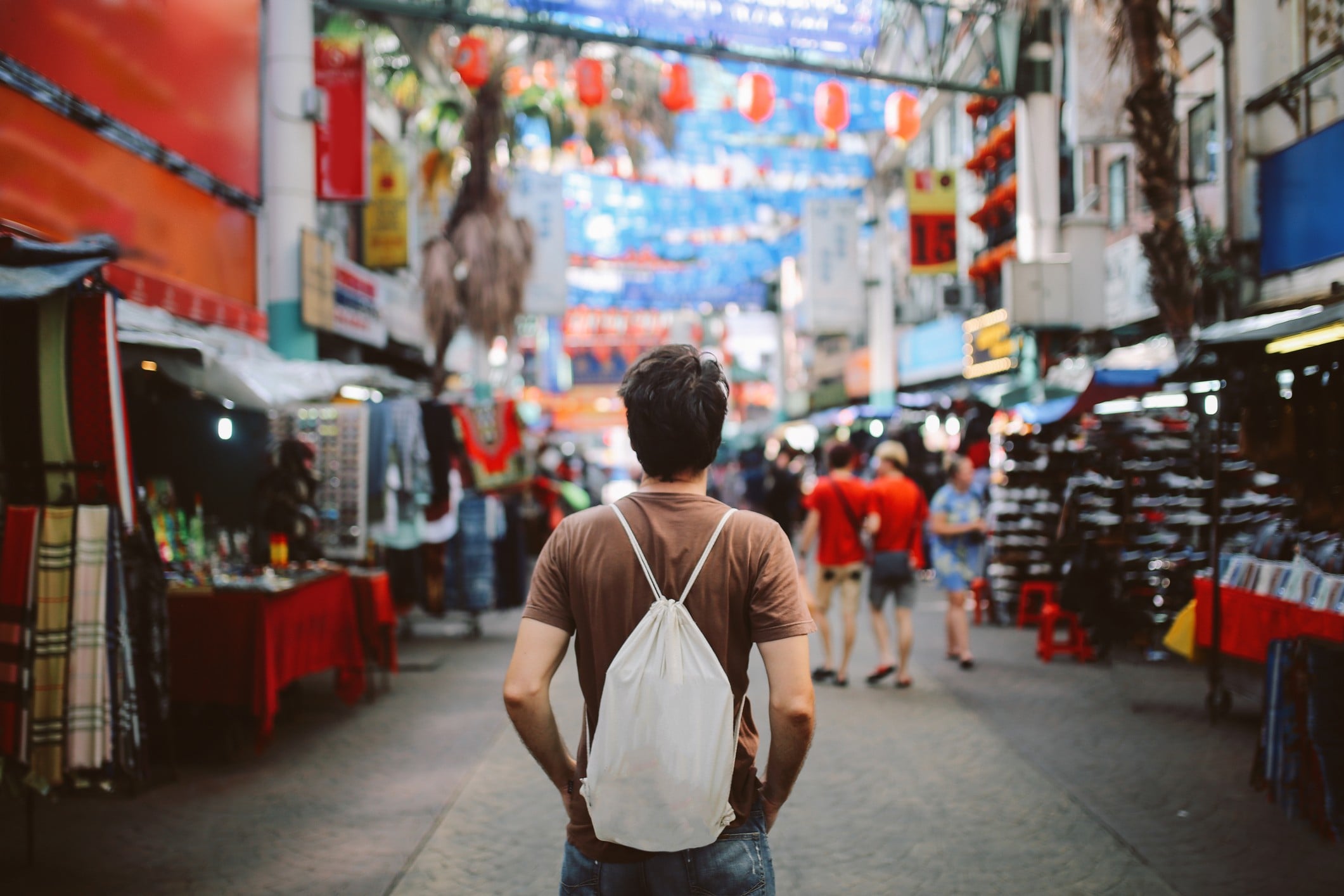 A person stands in a bustling market, facing colorful stalls and decorations, with many people in the background.