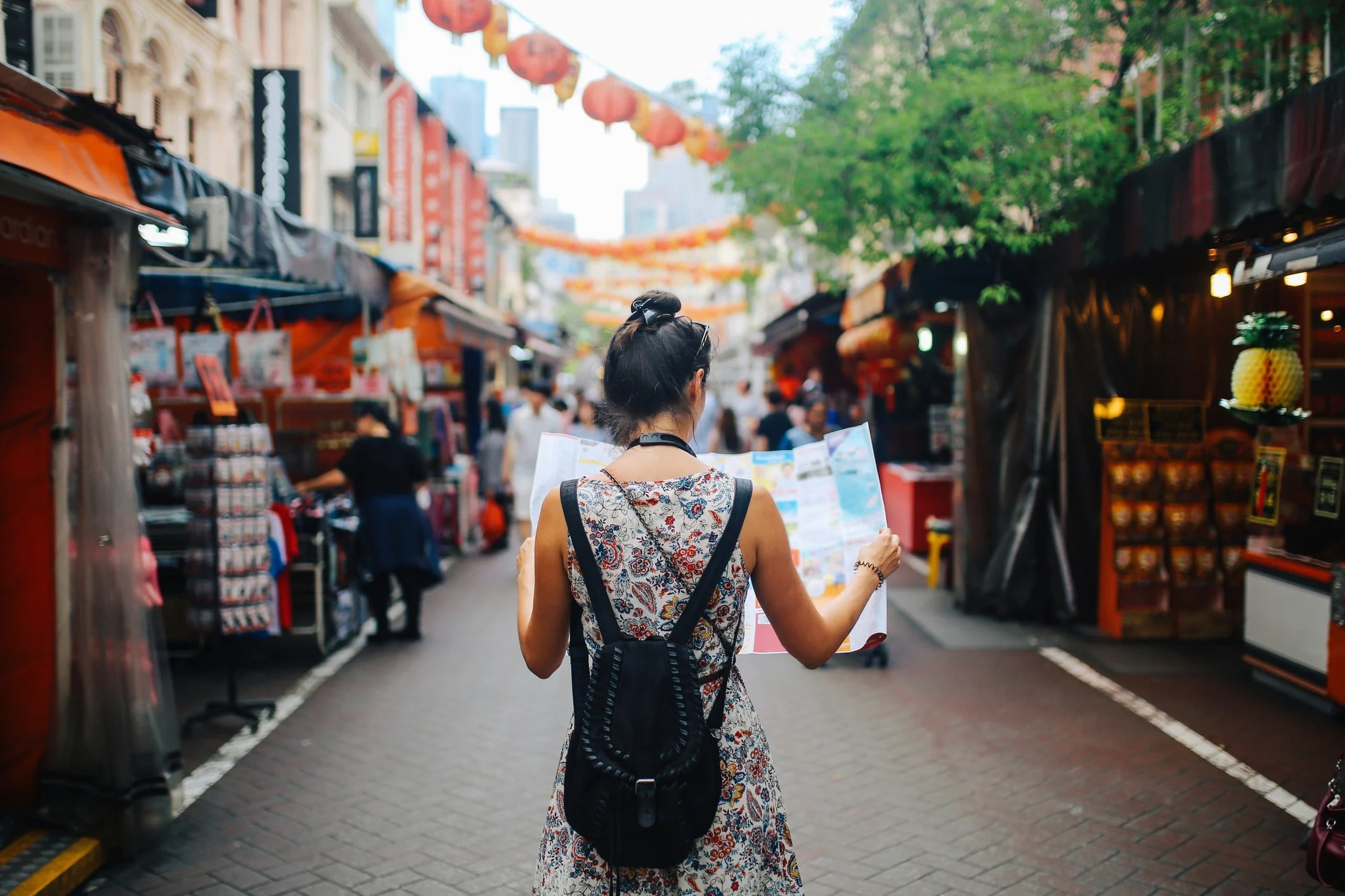 A woman stands in a market holding a map, surrounded by colorful stalls and hanging lanterns.